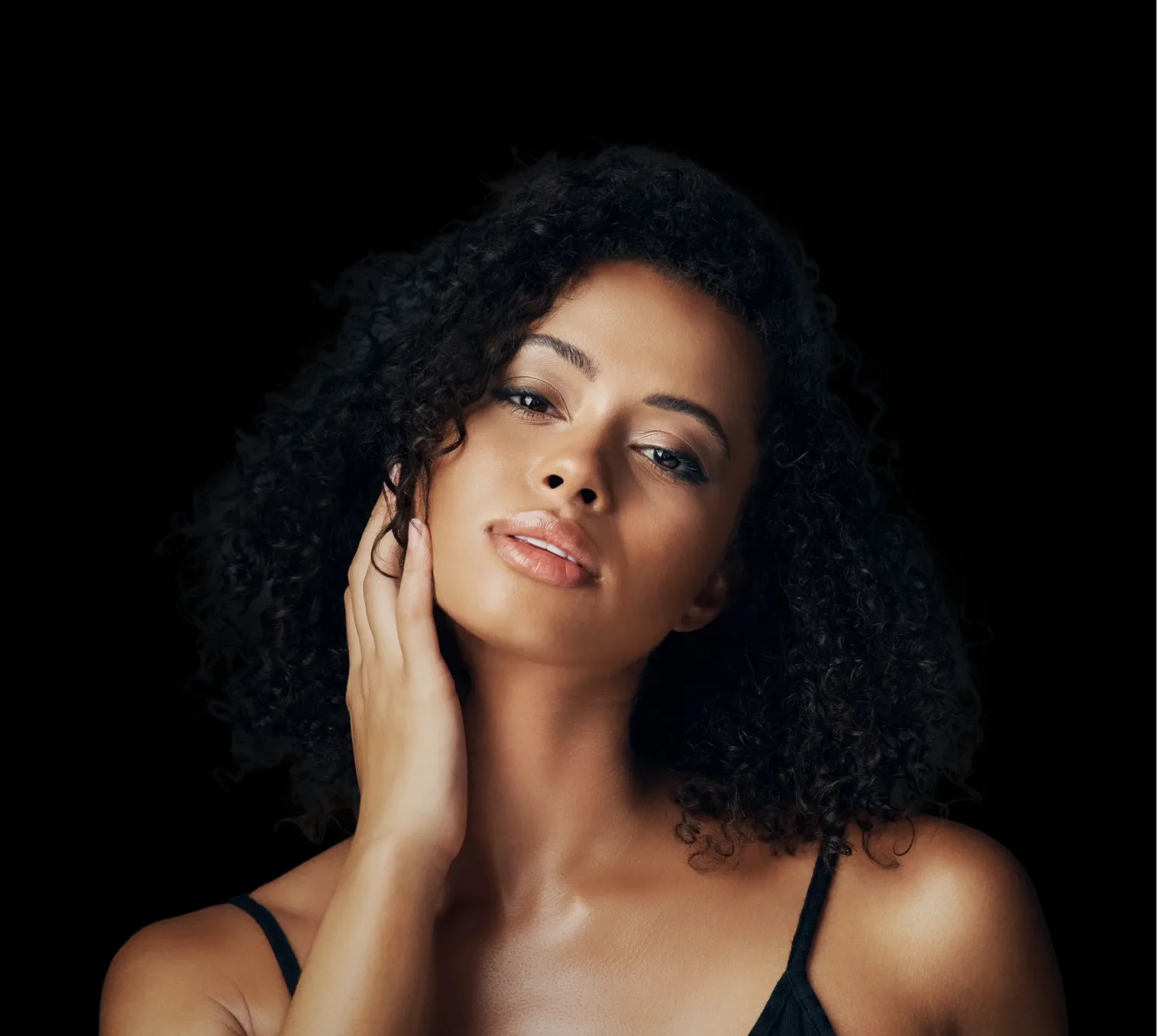 Woman with long curly dark hair touching her face against black background