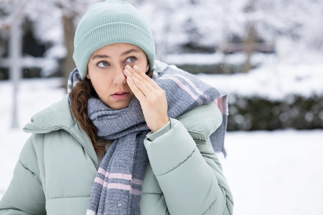 Woman in winter coat and beanie touching her face in pain outdoors in snow