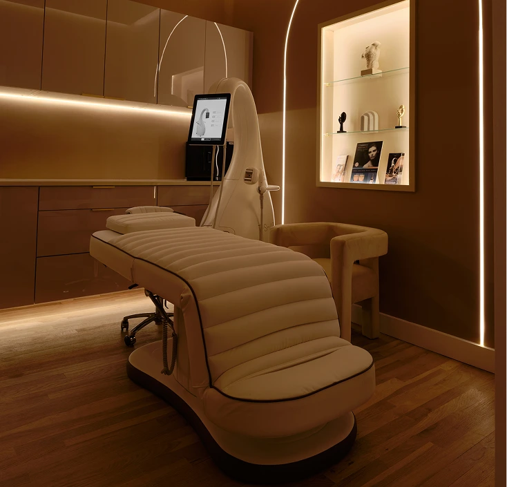 Modern treatment room at La Reservé with beige padded treatment bed, overhead device, warm lighting, and decorative shelf
