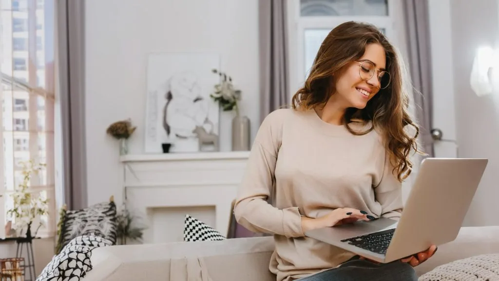 Woman in a bright home office holding a laptop, smiling while looking at the screen