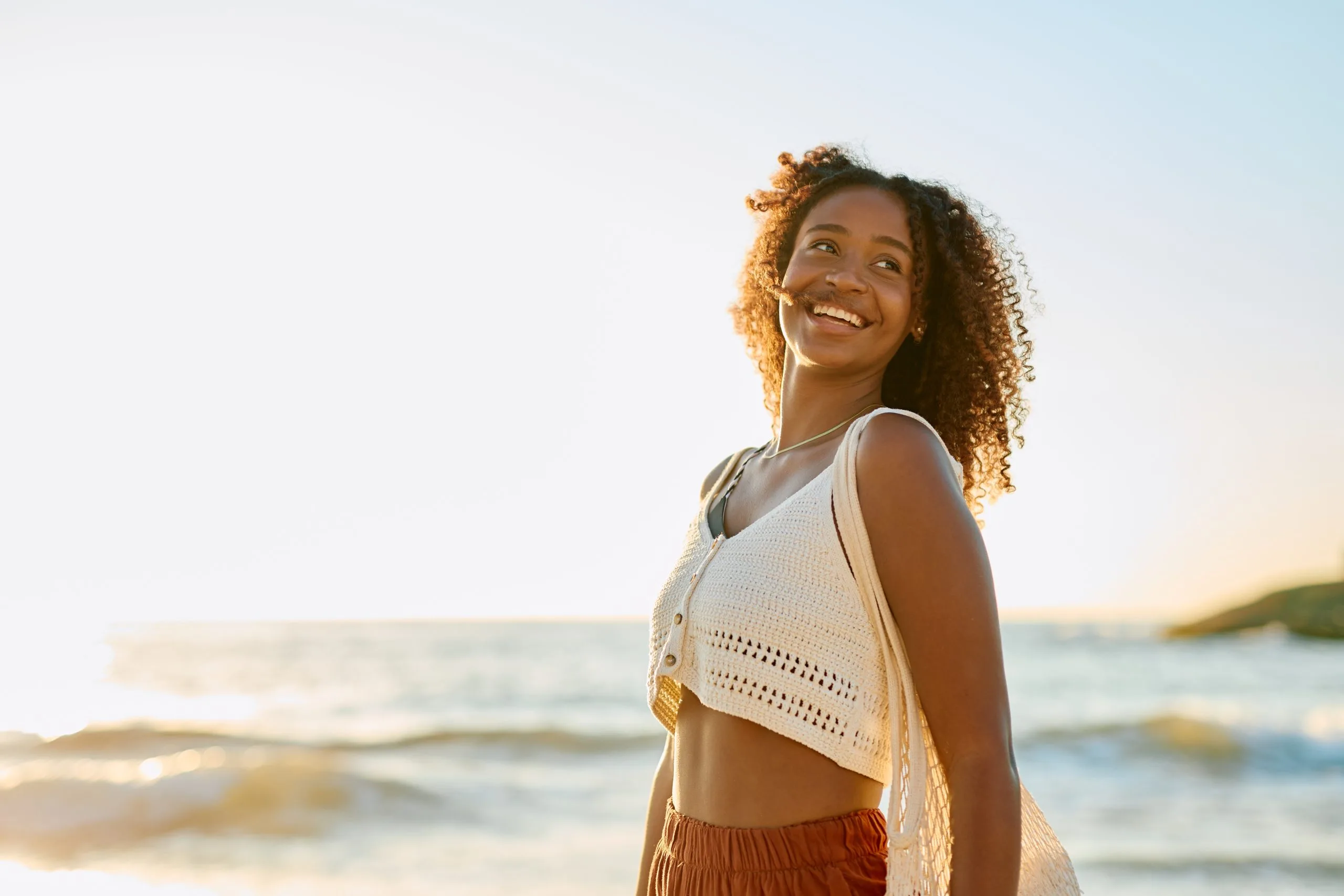 Woman with curly hair smiling while standing on a beach at sunset