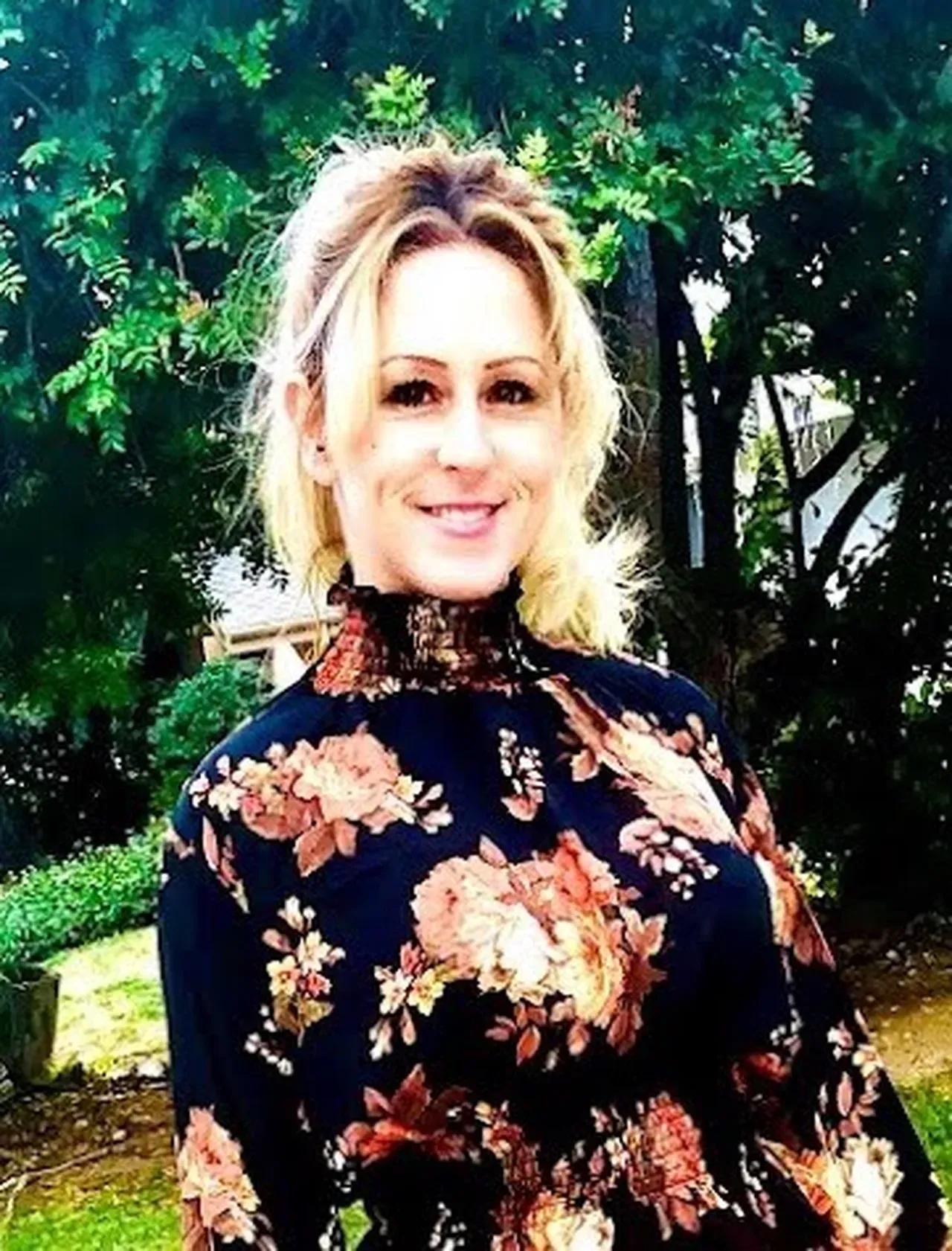 Woman with blonde hair smiling in front of green foliage wearing navy floral top