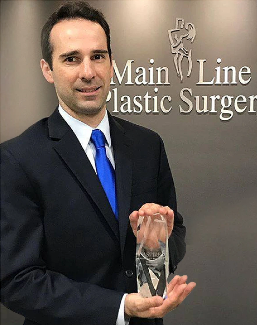 Man in dark suit and blue tie holding award with Main Line Plastic Surgery logo on wall behind