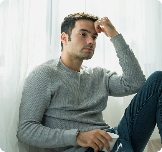 Man in gray sweater sitting indoors, hand raised to head, looking thoughtful