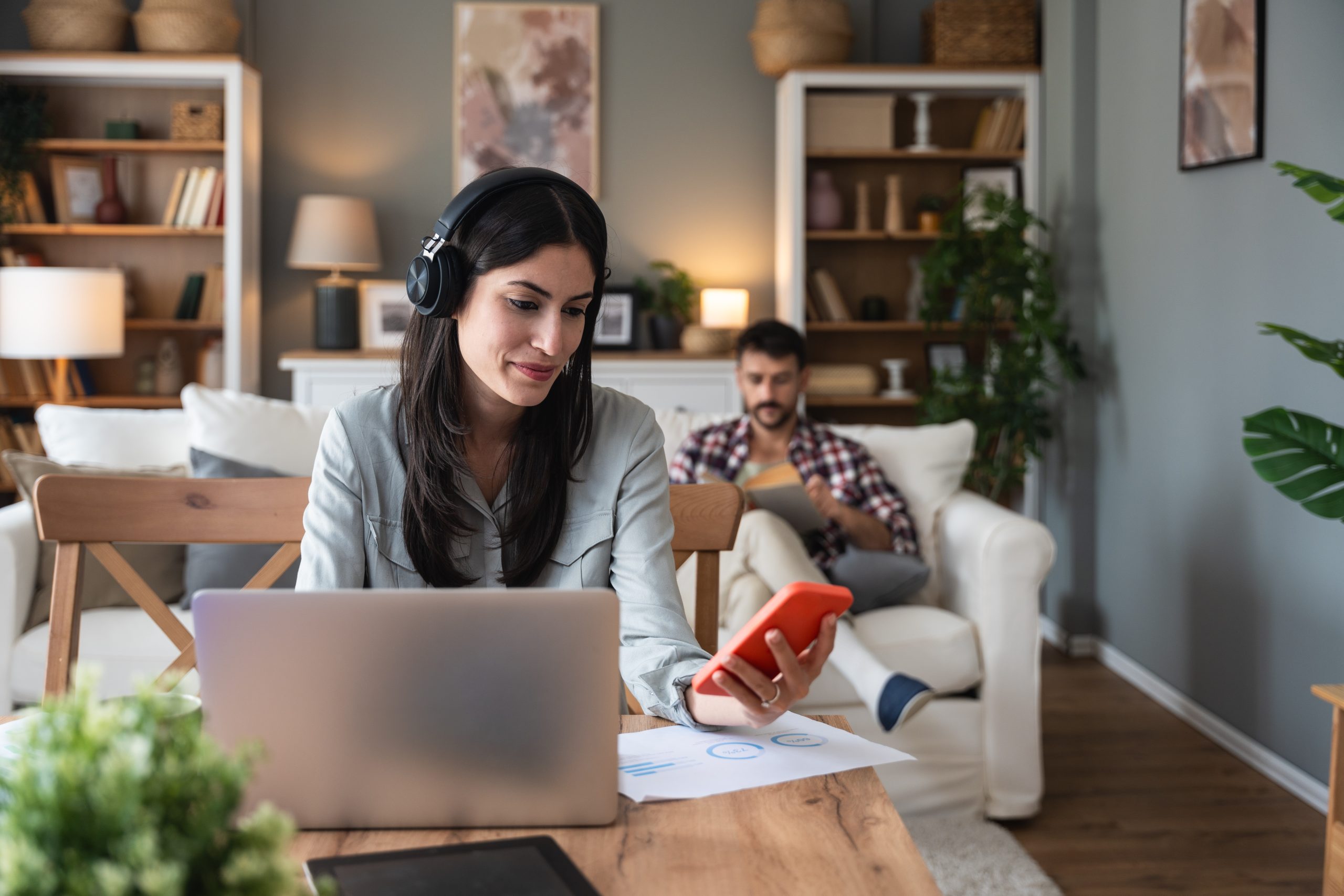 Woman wearing headphones smiles at laptop while holding a red mug, with a man using a tablet on a couch behind her