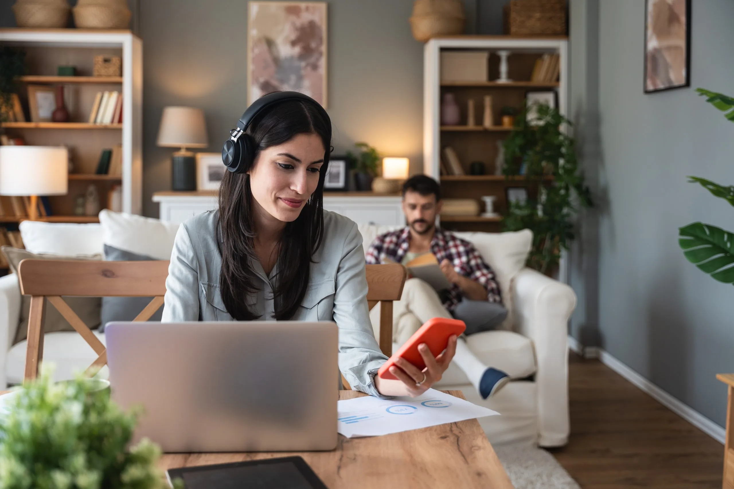 Woman wearing headphones smiles at laptop while holding a red mug, with a man using a tablet on a couch behind her