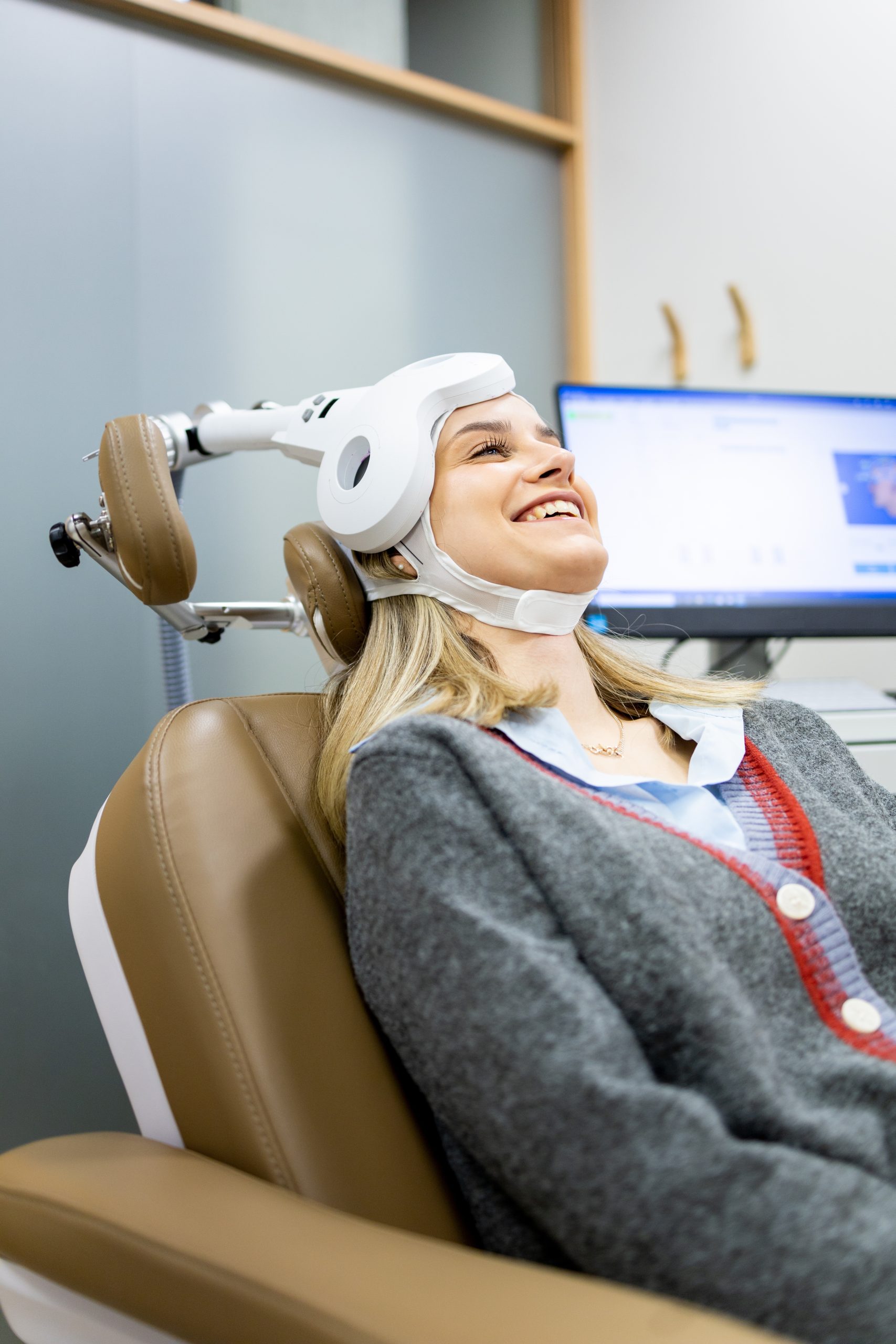 Patient reclines in dental chair wearing white head apparatus while smiling, with monitor visible in background