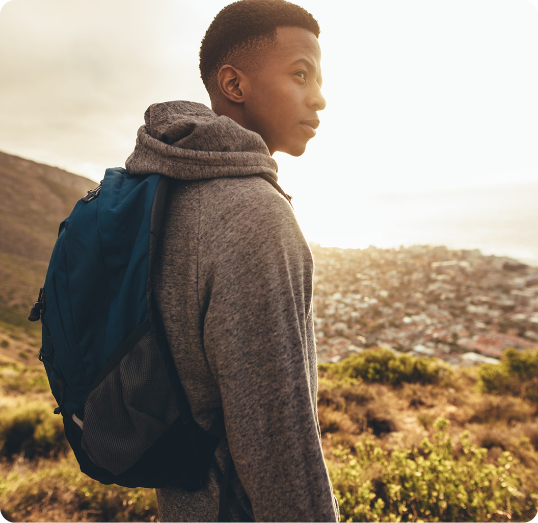 Young man wearing blue backpack and gray hoodie looking over shoulder at rural hillside landscape