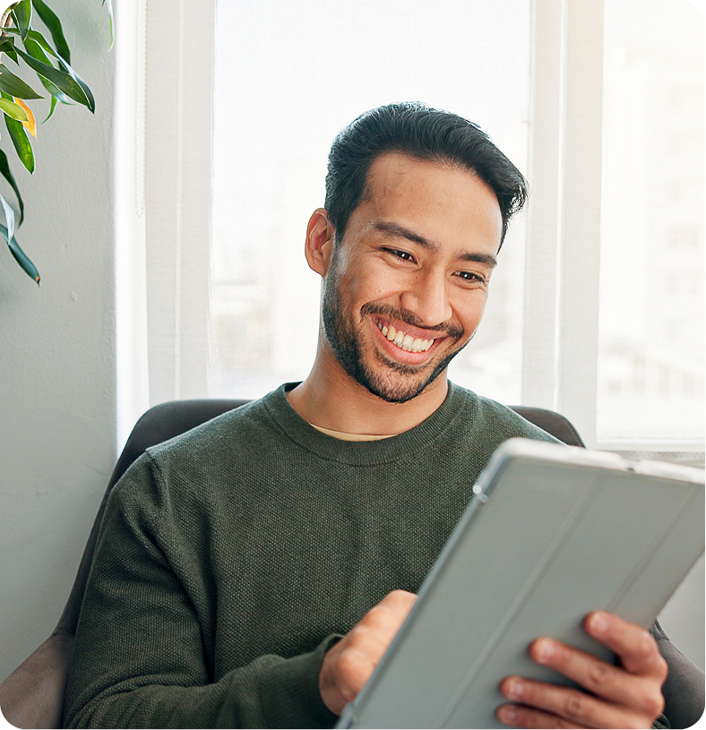 Man smiling while holding a tablet device by a bright window with a potted plant