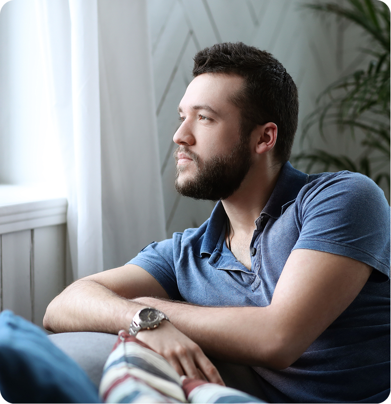 Man in blue shirt sitting indoors by a window, looking thoughtful