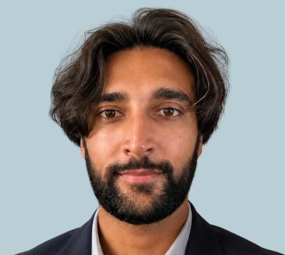 Man with dark hair and beard wearing dark blazer against light blue background