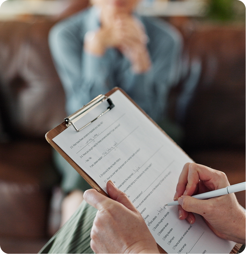 Clinician holding clipboard and pen while patient in light blue shirt sits on couch during psychiatric assessment
