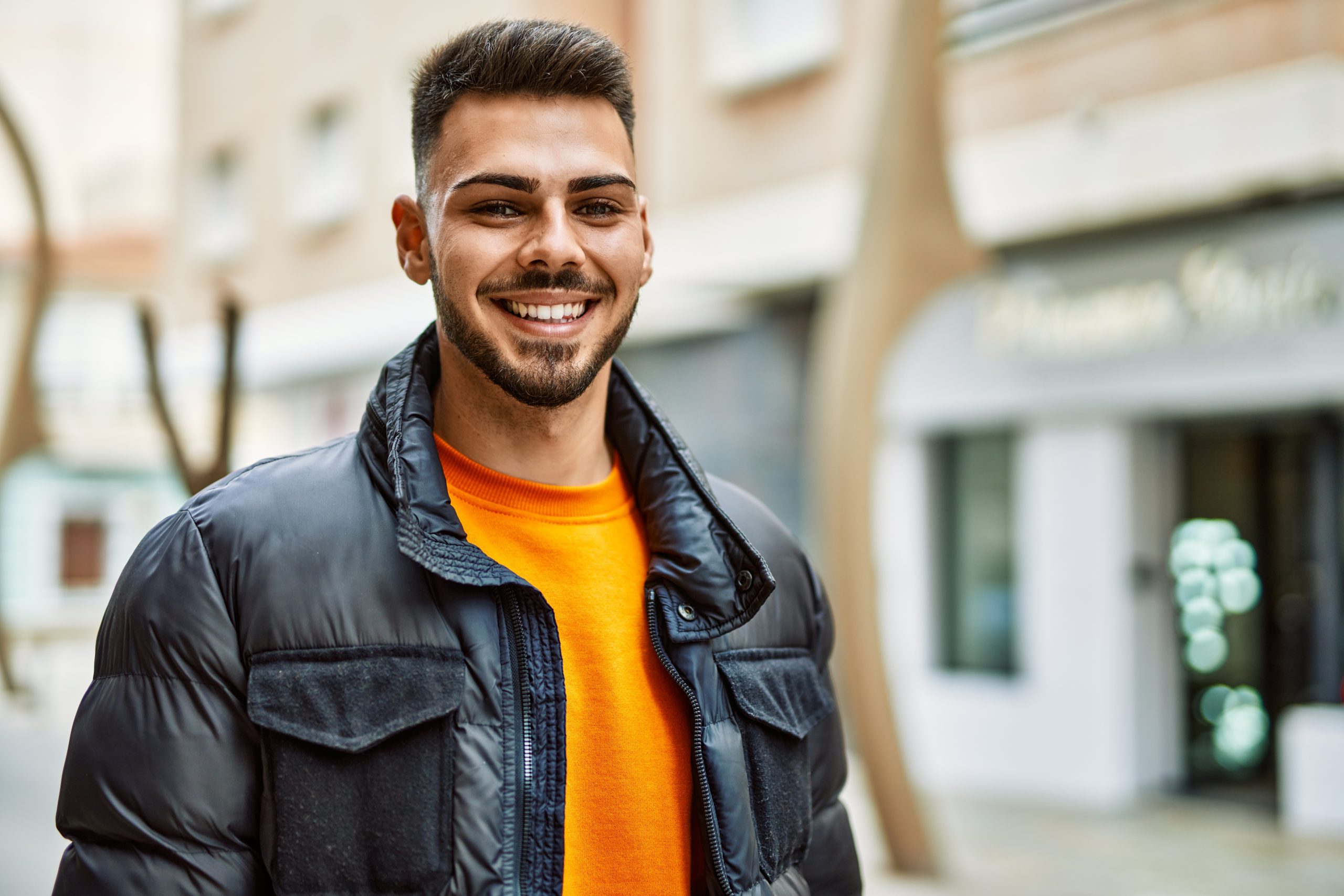 Man smiling wearing orange shirt and black jacket on city street