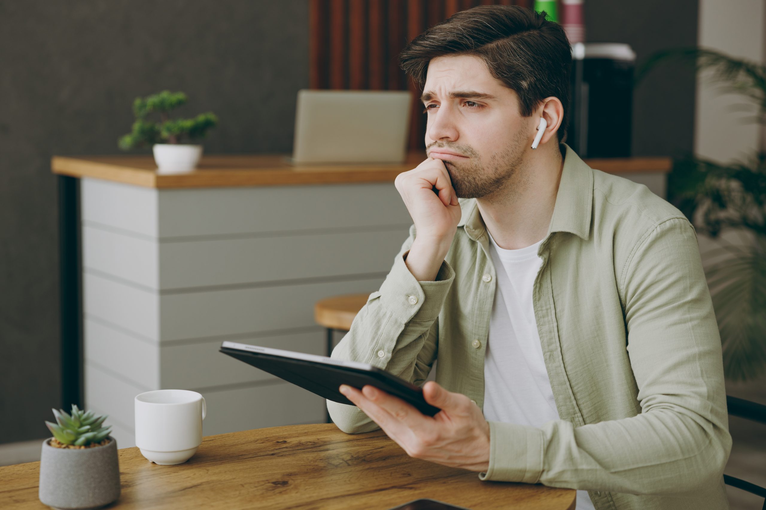 Man in sage green shirt holding tablet at desk, looking contemplative with earbuds