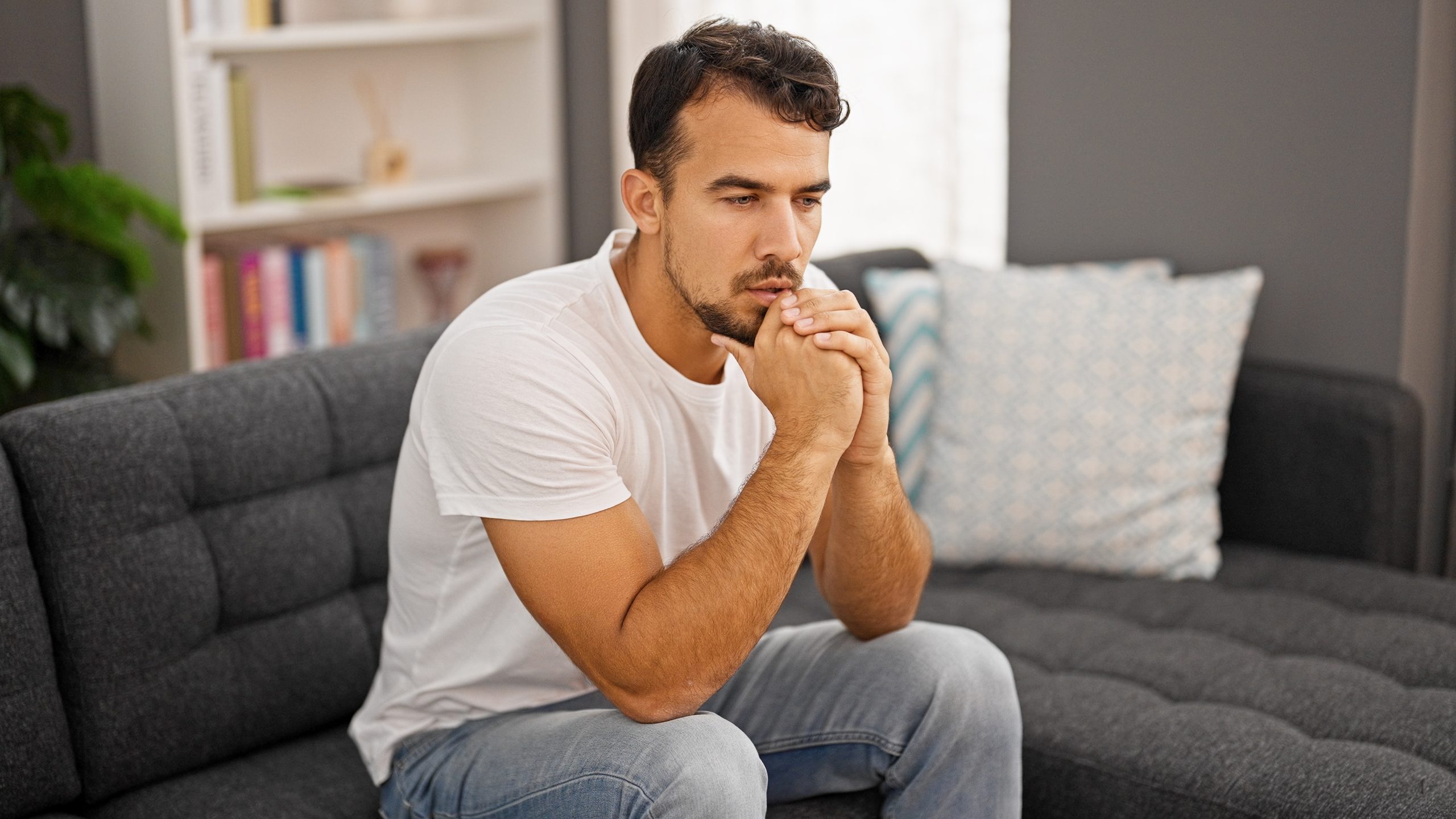 Man in white shirt sitting on gray couch with hand to chin, looking thoughtful