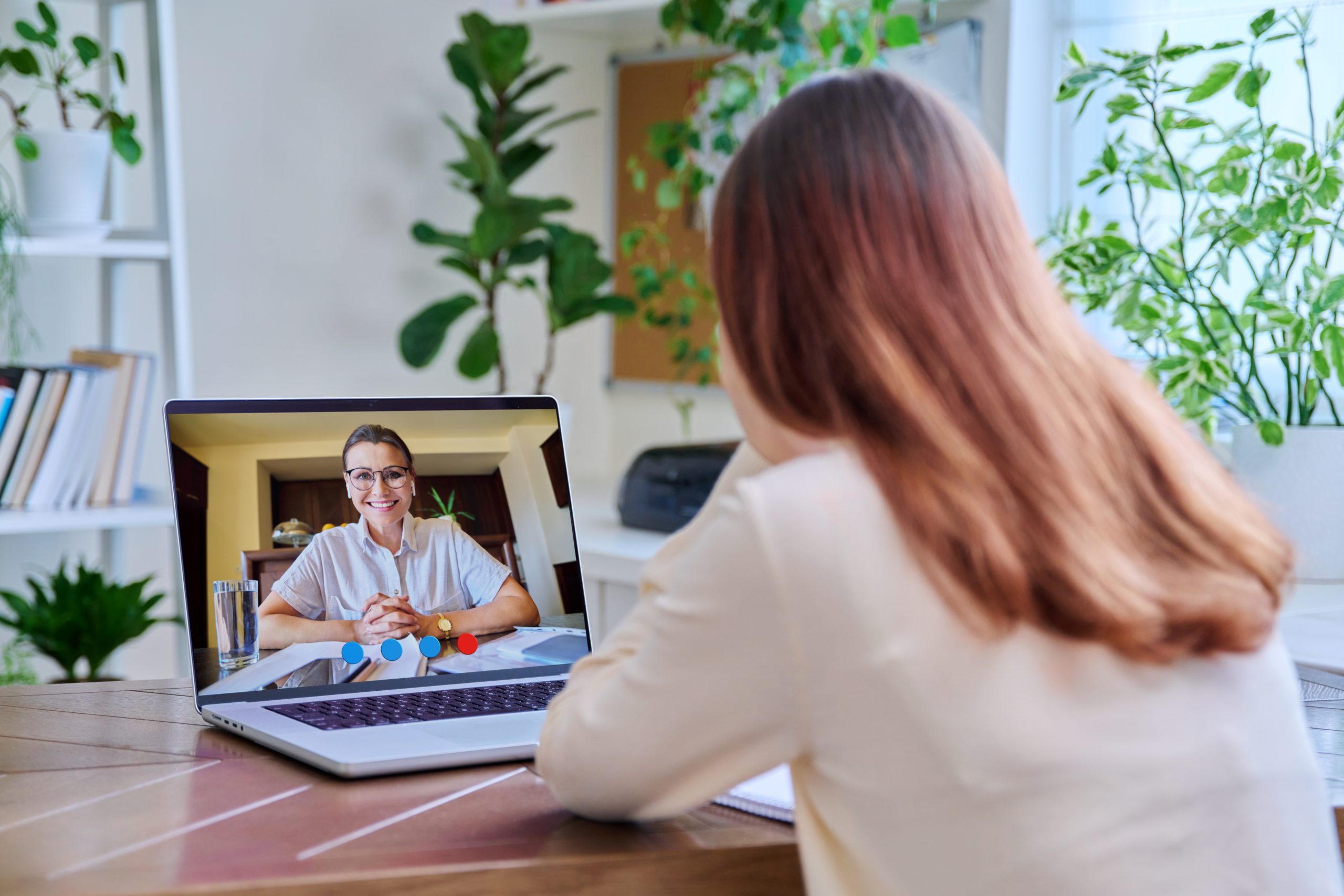 Woman viewed from behind during video call with man on laptop screen in plant-filled office