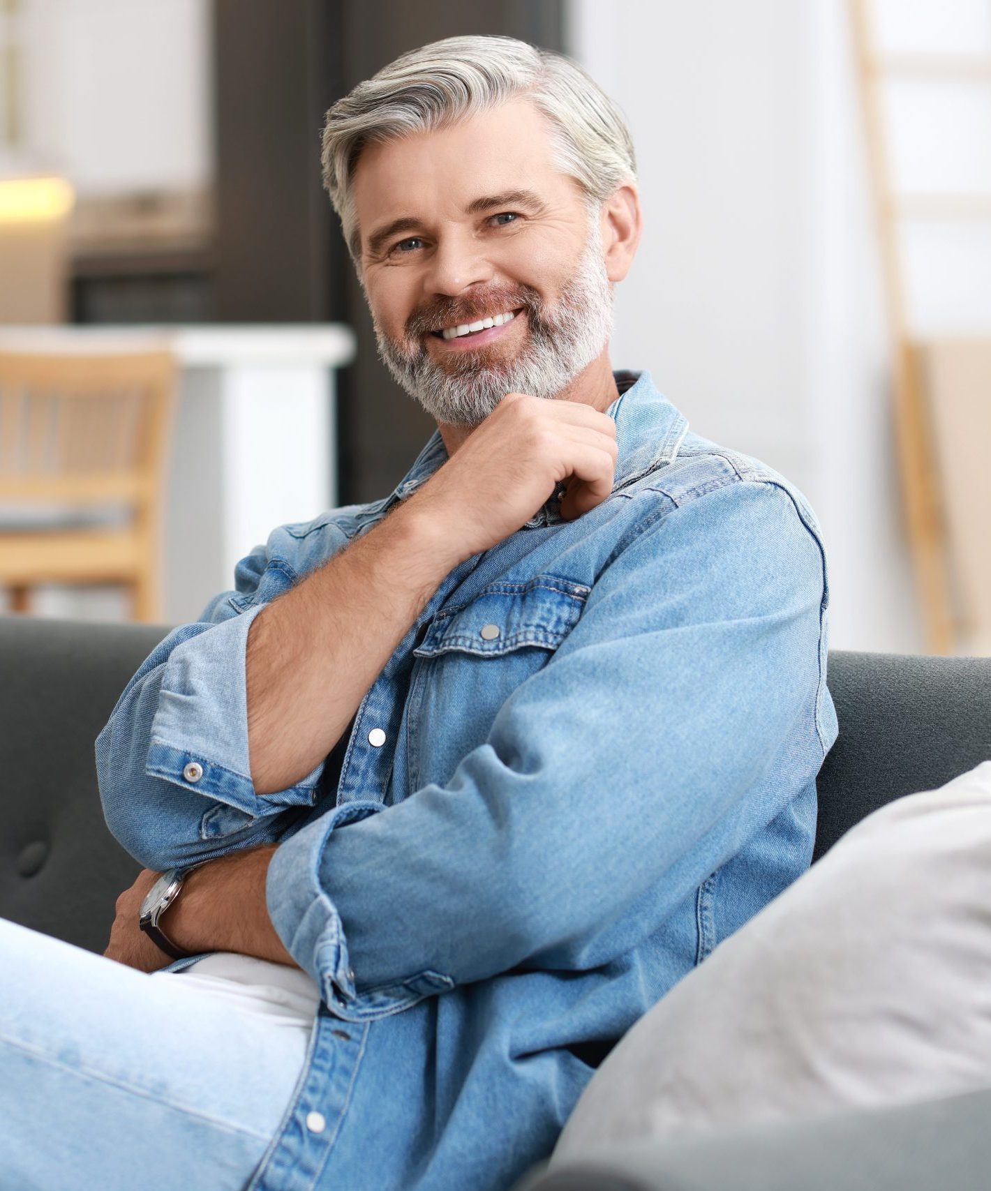 Middle-aged man with gray beard smiling at camera, wearing denim shirt, sitting indoors