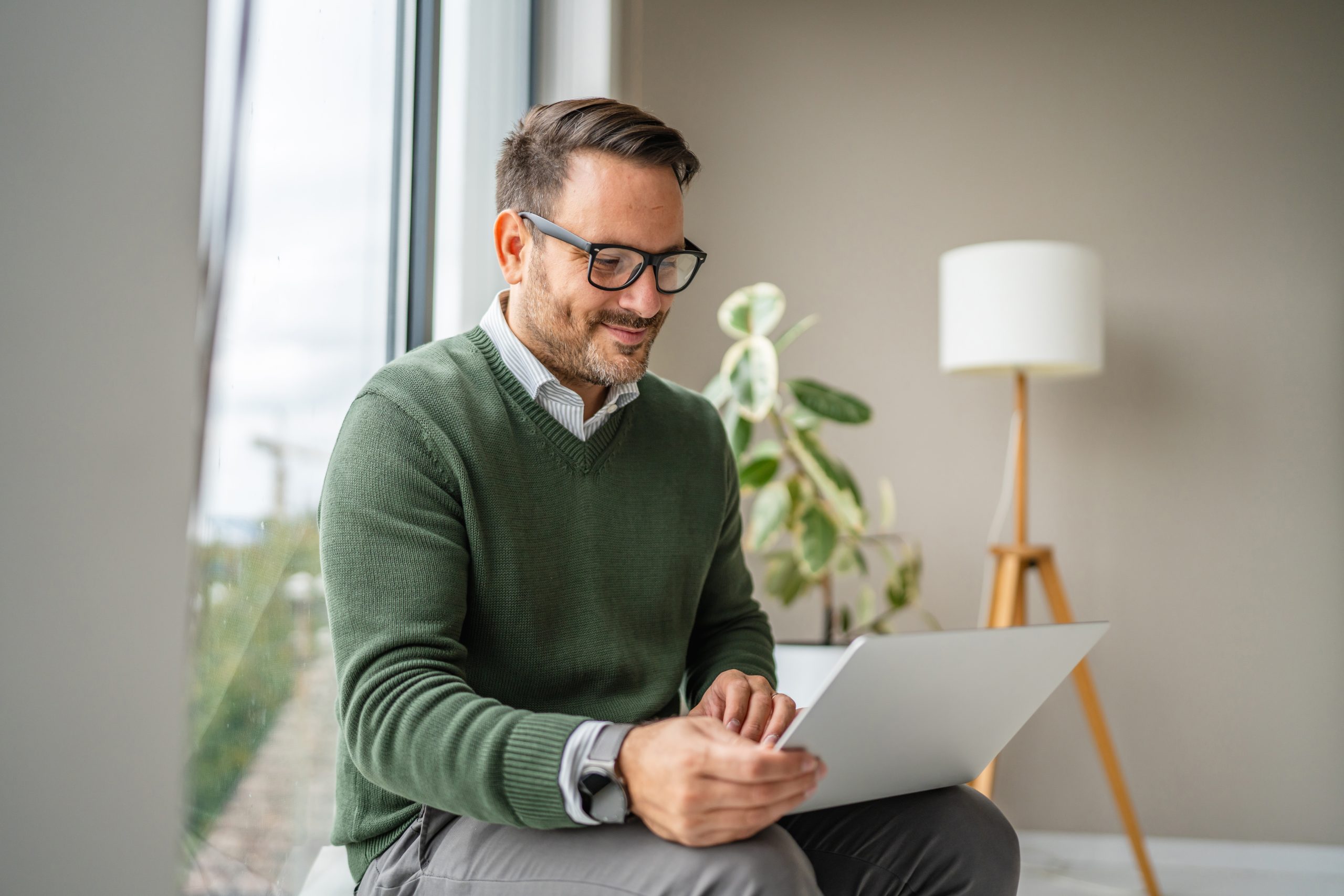 Man in green sweater using laptop by window with potted plant and lamp