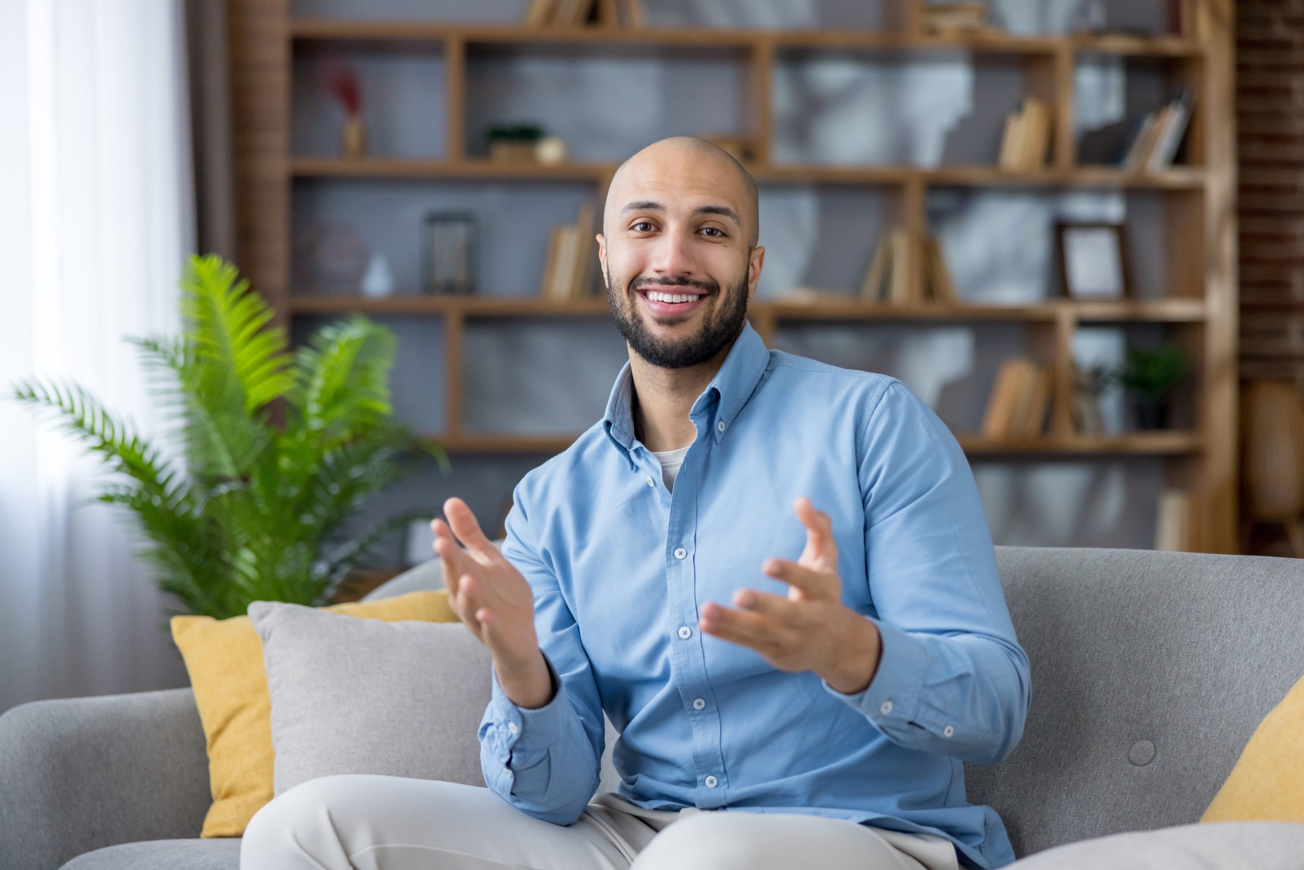 Man in light blue shirt gesturing while speaking, seated on couch with shelving and plant behind him