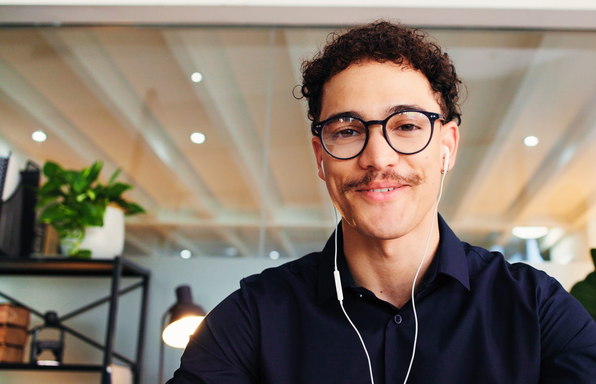 Man wearing glasses and headphones smiling in modern office setting with plants