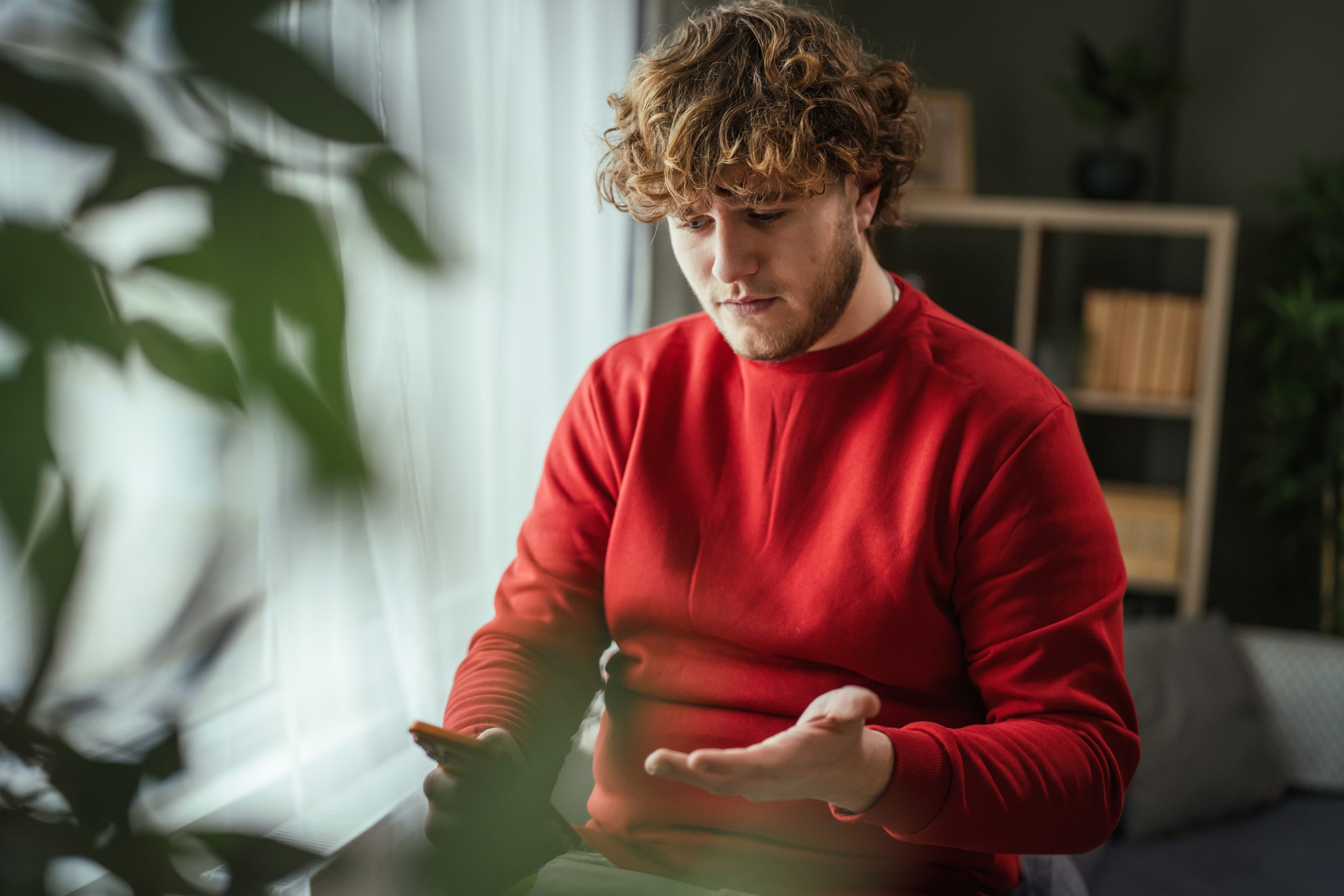 Man in red sweater looking at smartphone indoors with plant and shelving visible