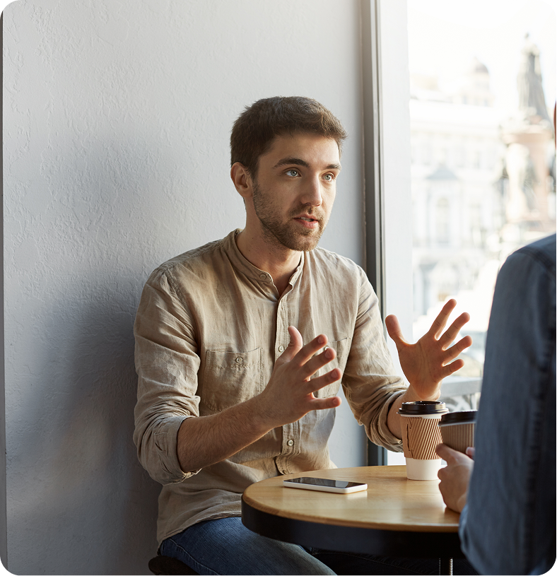 Man in beige shirt gesturing while speaking with another person during a therapy session by a window