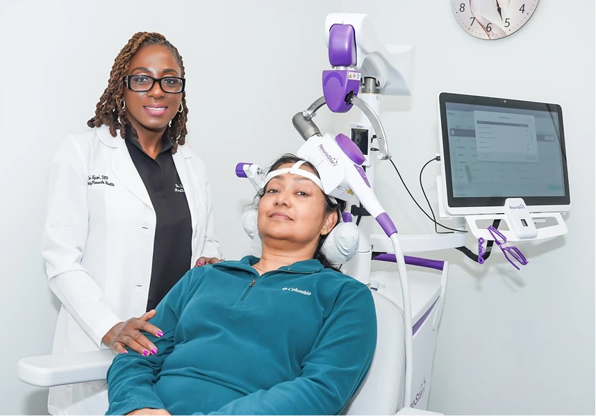 Clinician in white coat standing beside patient receiving TMS therapy with purple-and-white headpiece and monitor display