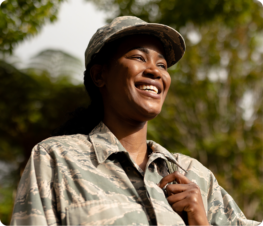 Service member in camouflage uniform smiling outdoors with trees in background