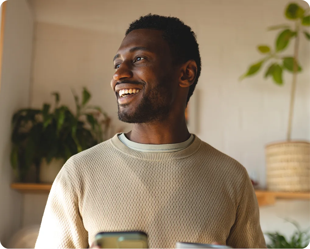 Man smiling in a bright room with potted plants, holding a phone