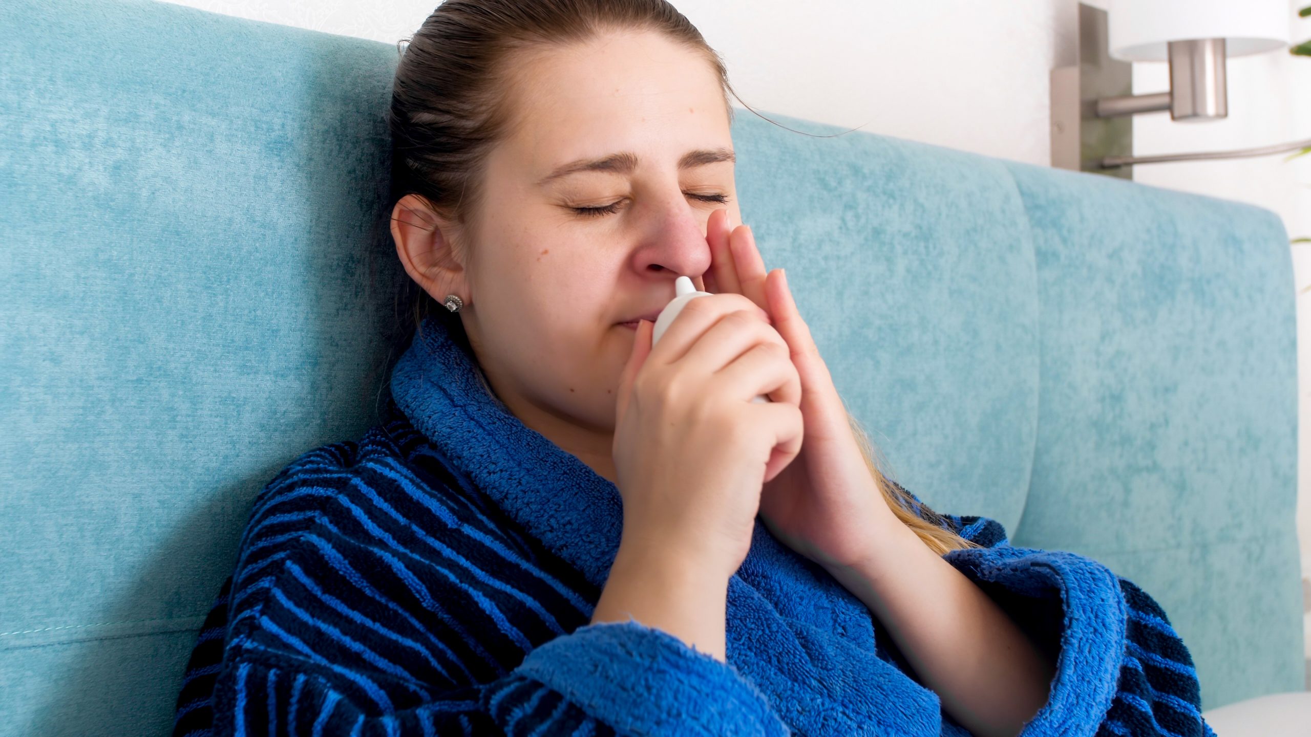 Woman on teal couch wrapped in blue blanket coughing into her hand