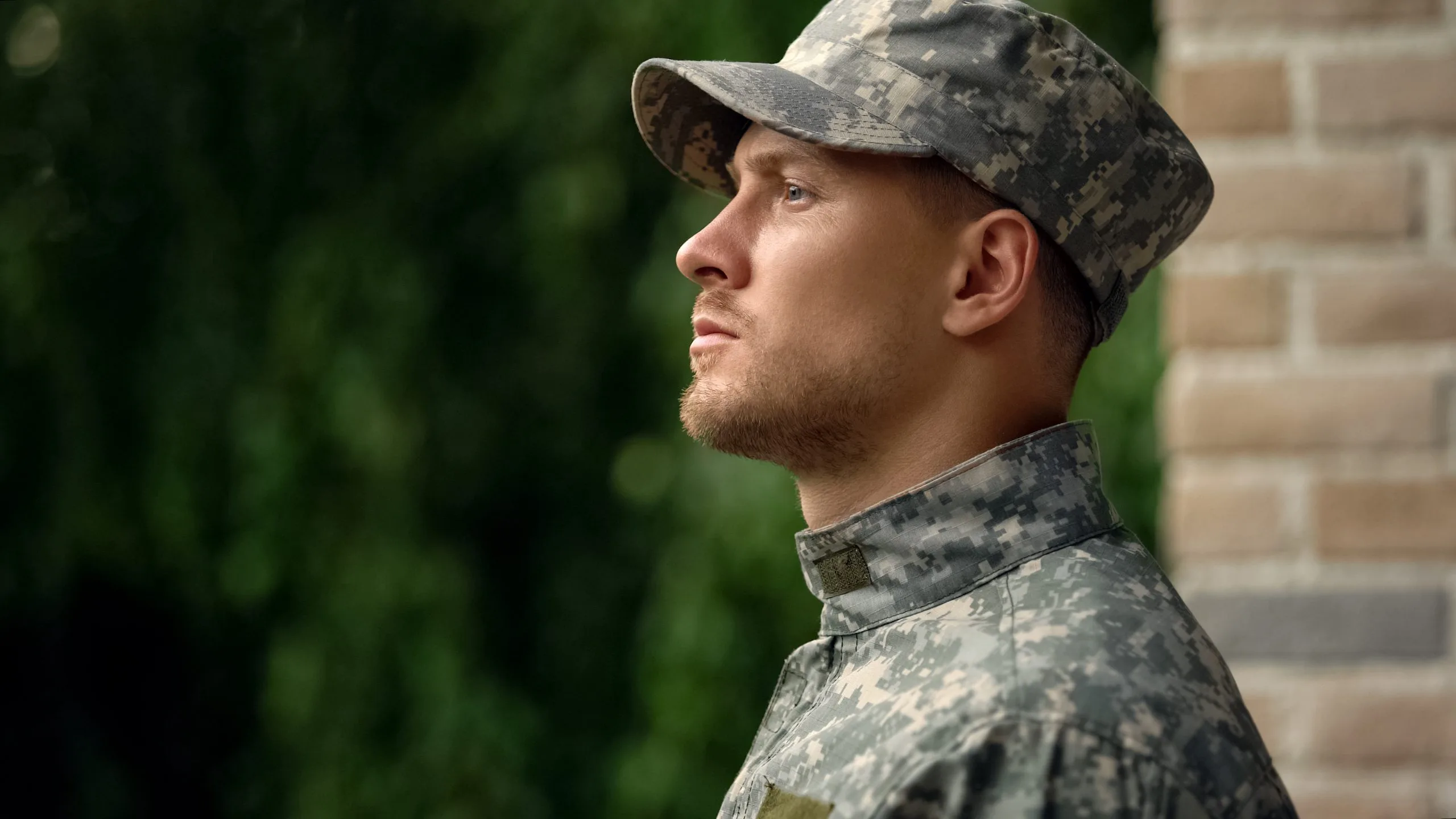 Male service member in digital camouflage uniform and cap looking to the side outdoors