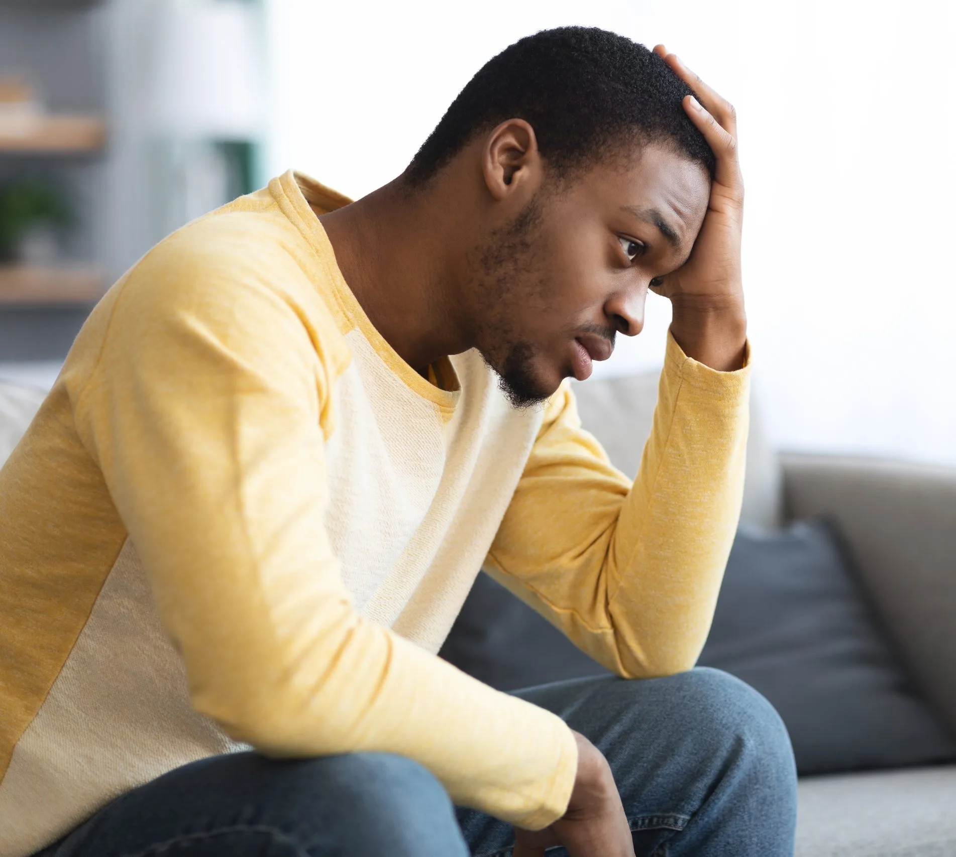 Man in yellow shirt sitting on couch with head in hand, looking troubled