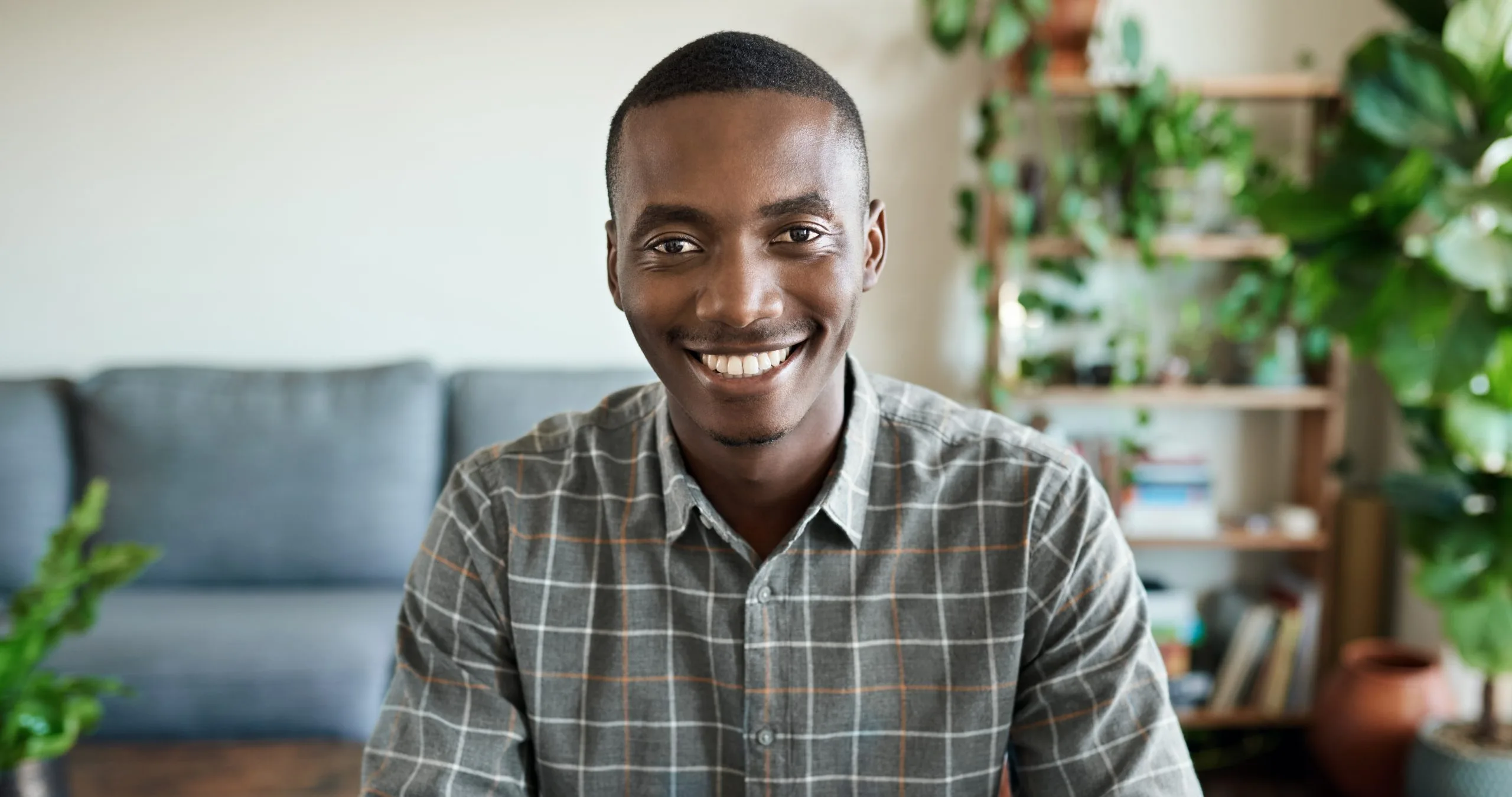 Smiling man in checkered shirt sitting on couch with potted plants behind