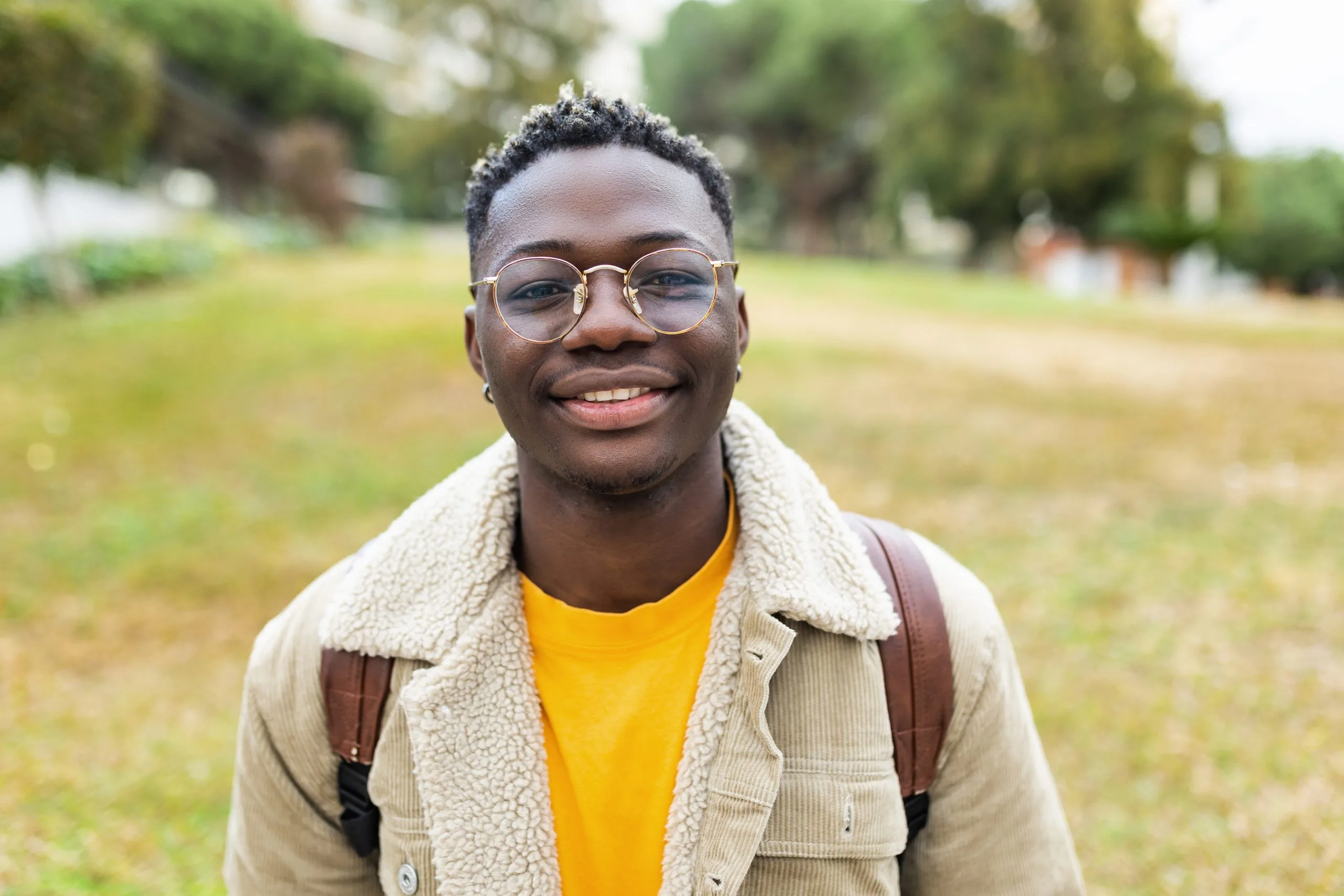 Man smiling at camera outdoors wearing glasses, yellow shirt, and tan jacket with backpack