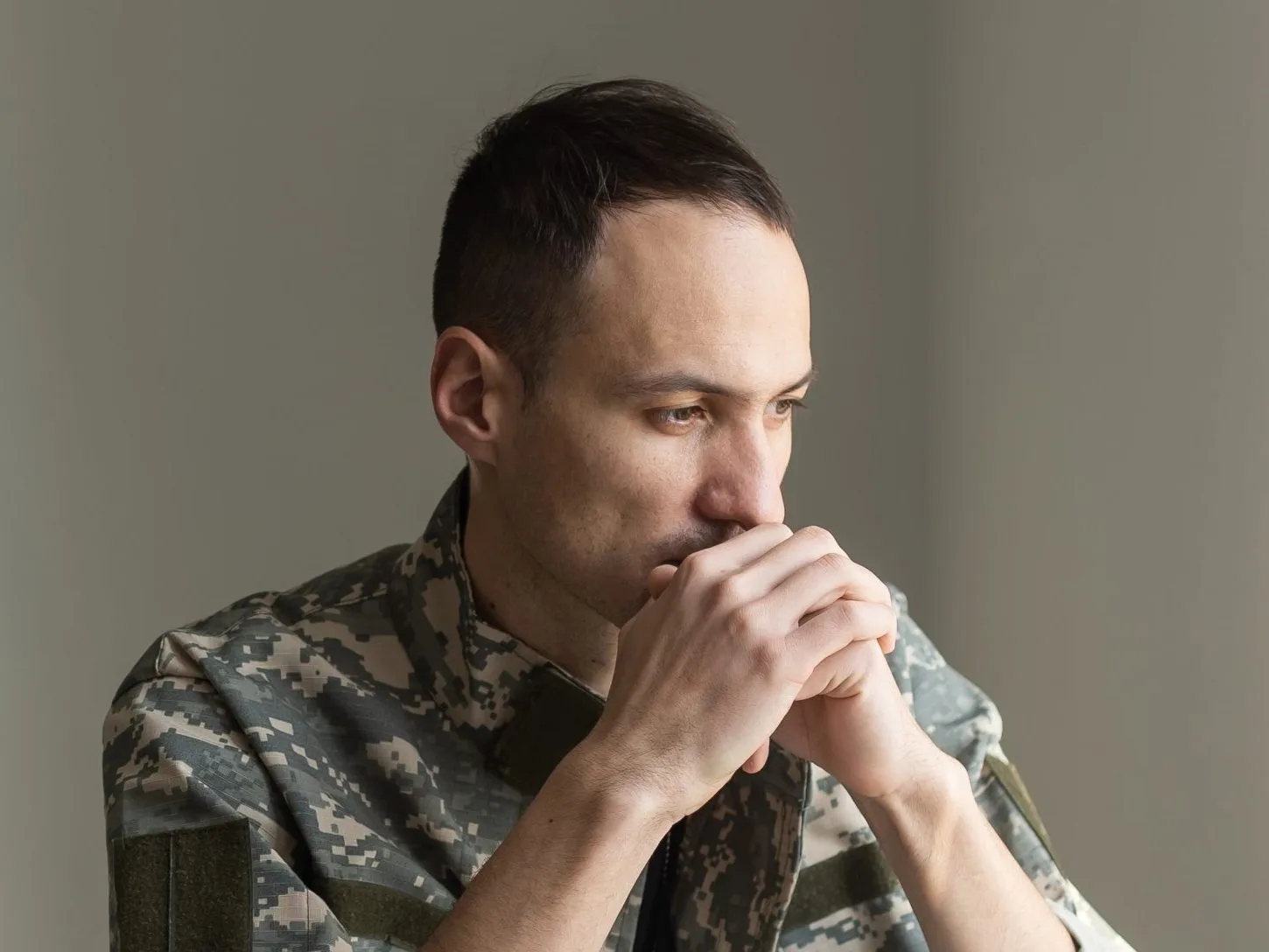 Service member in camouflage uniform sitting with hands clasped, looking thoughtful