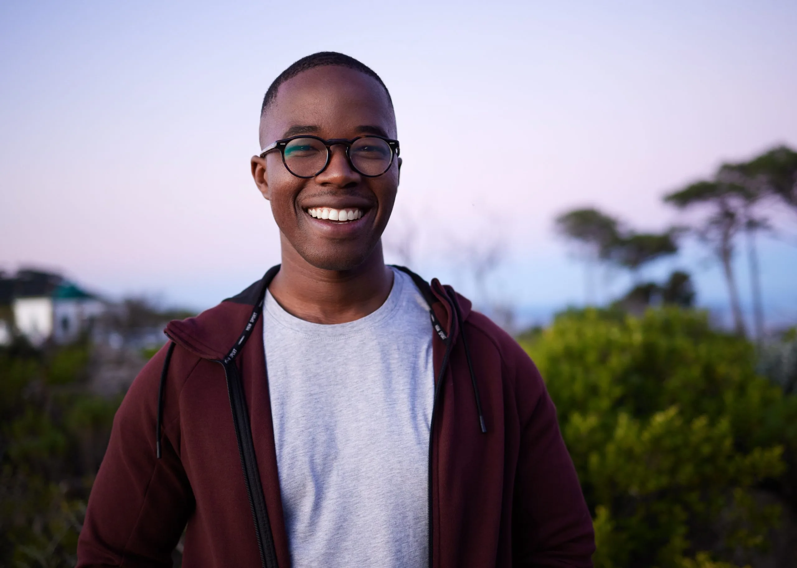 Smiling man wearing glasses and maroon hoodie outdoors at sunset with trees in background
