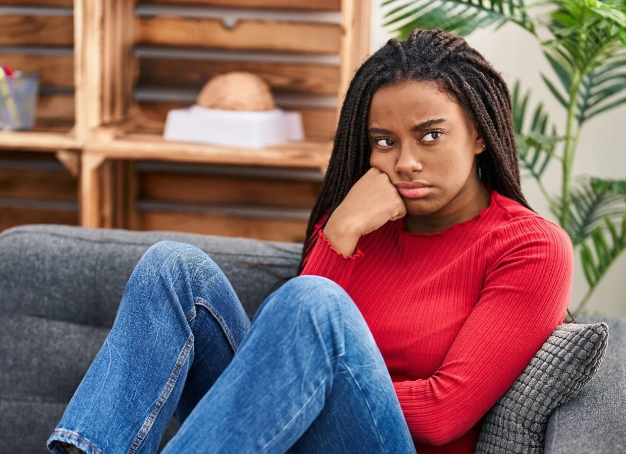 Woman in red sweater sitting on couch looking concerned with blurred figure in background
