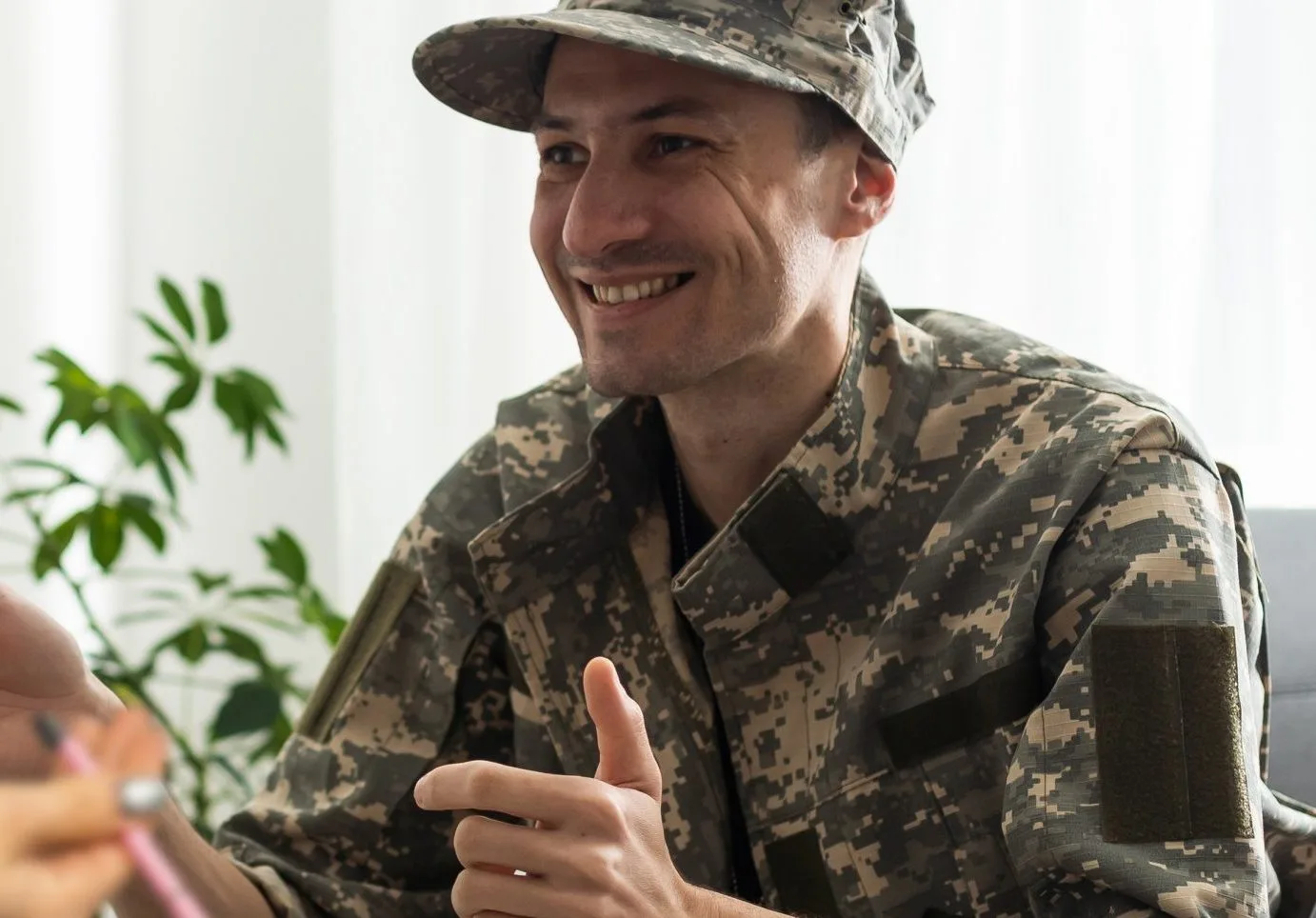 Smiling service member in digital camouflage uniform and cap gesturing while speaking