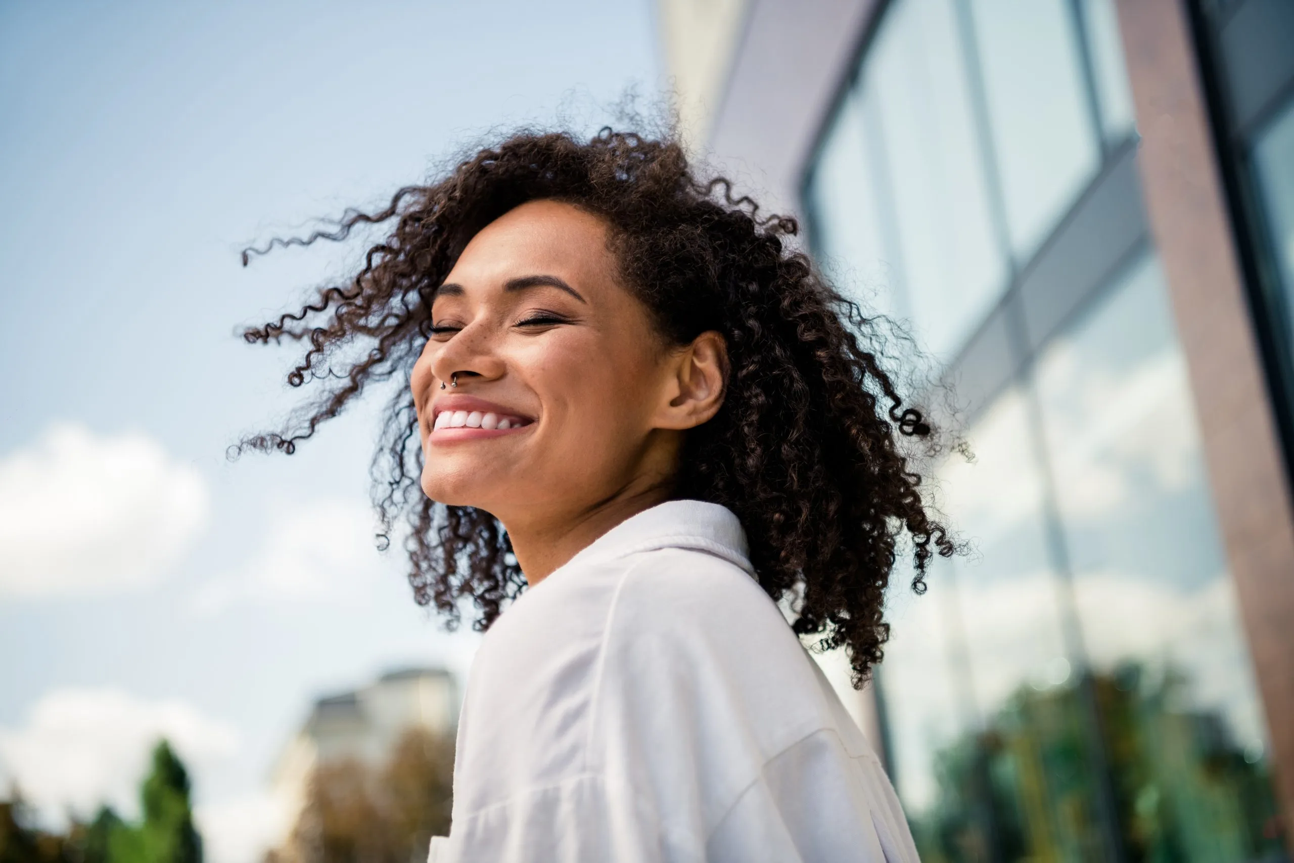 Woman with curly hair smiling outdoors against modern building backdrop