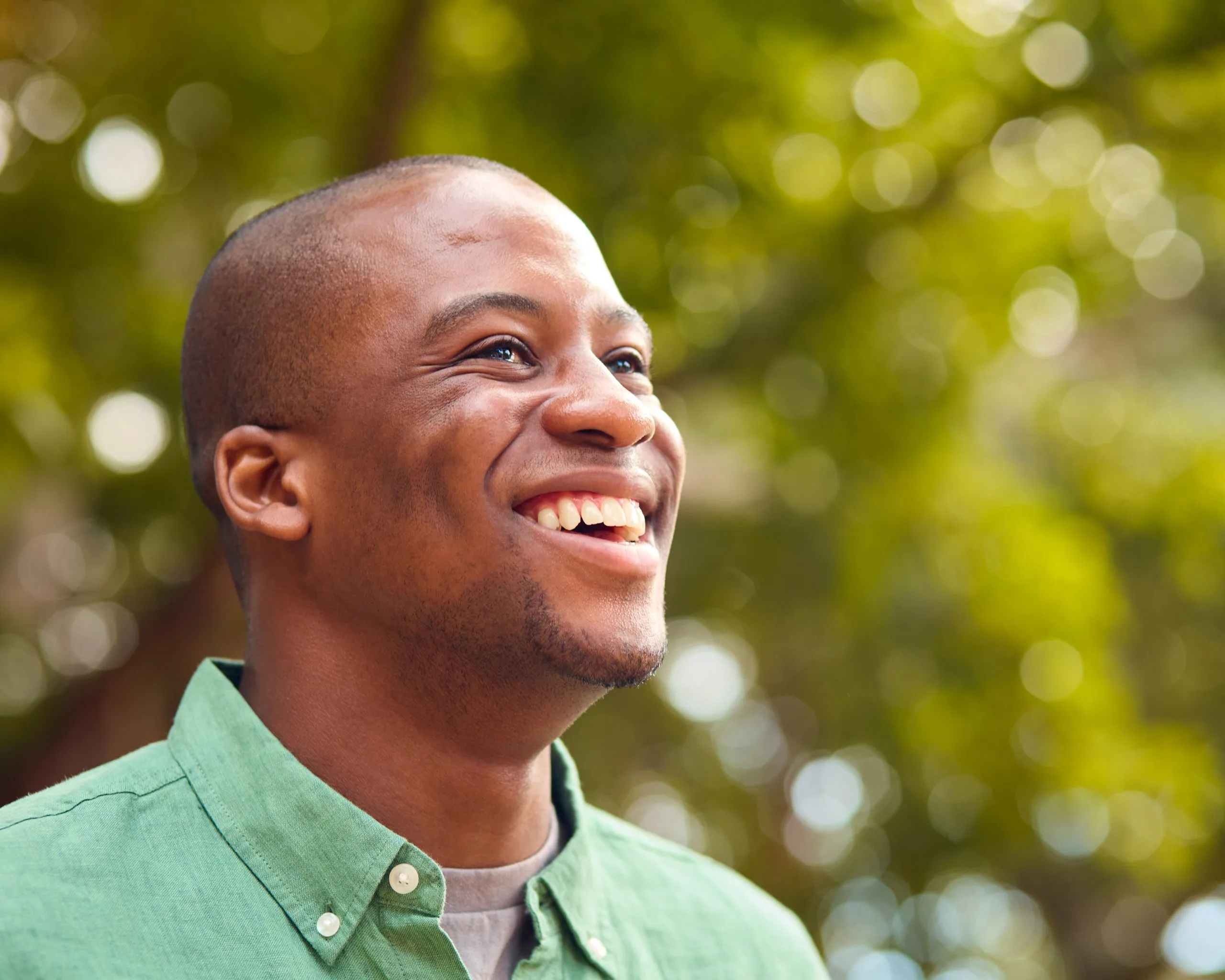 Man smiling outdoors in green button-up shirt with blurred green foliage background