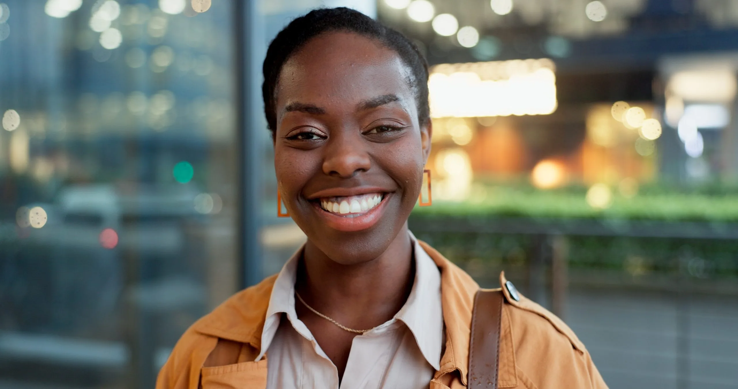 Smiling woman in orange jacket and white collar shirt against evening city lights and storefront