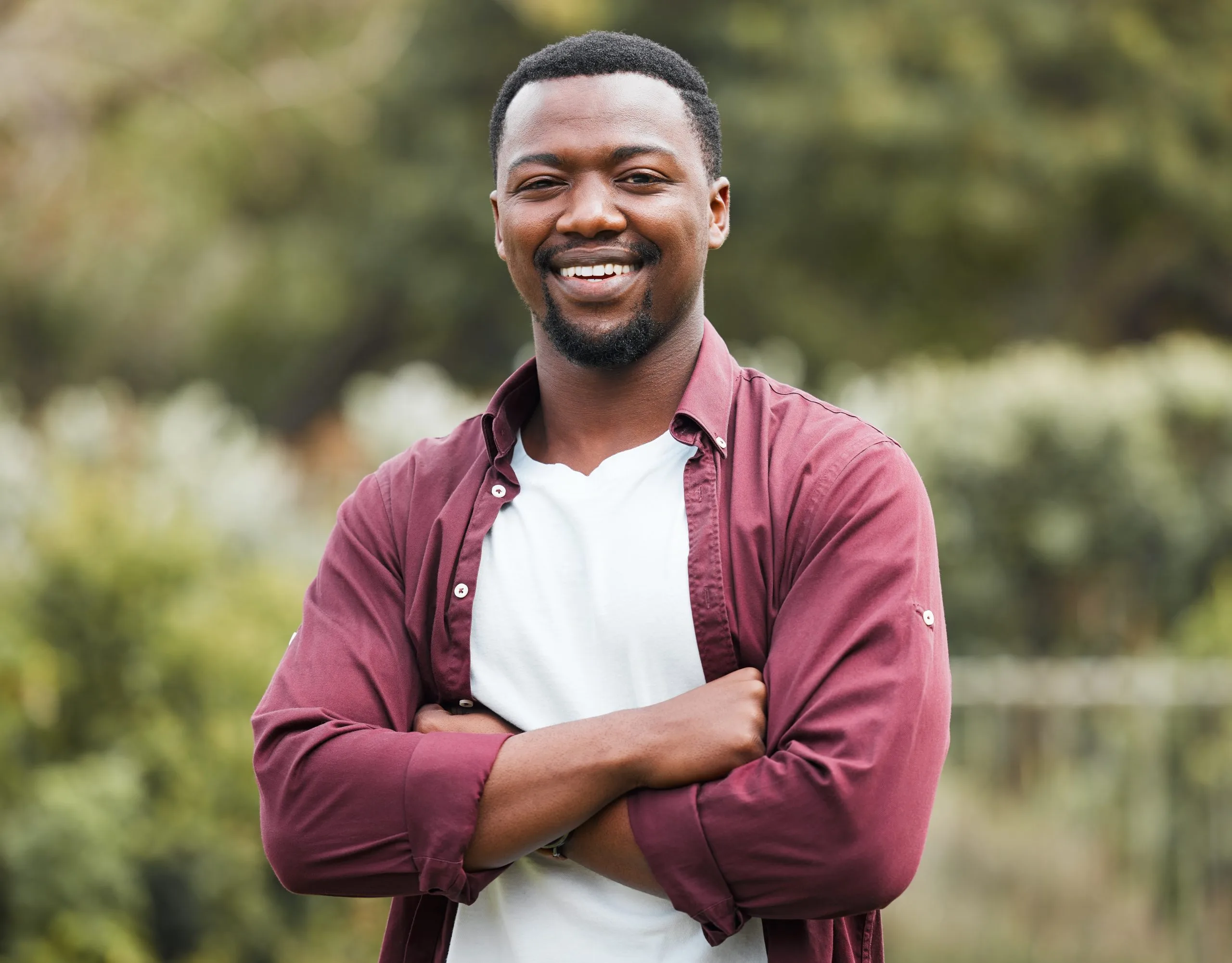 Man with arms crossed smiling at camera outdoors in garden wearing maroon shirt over white tee
