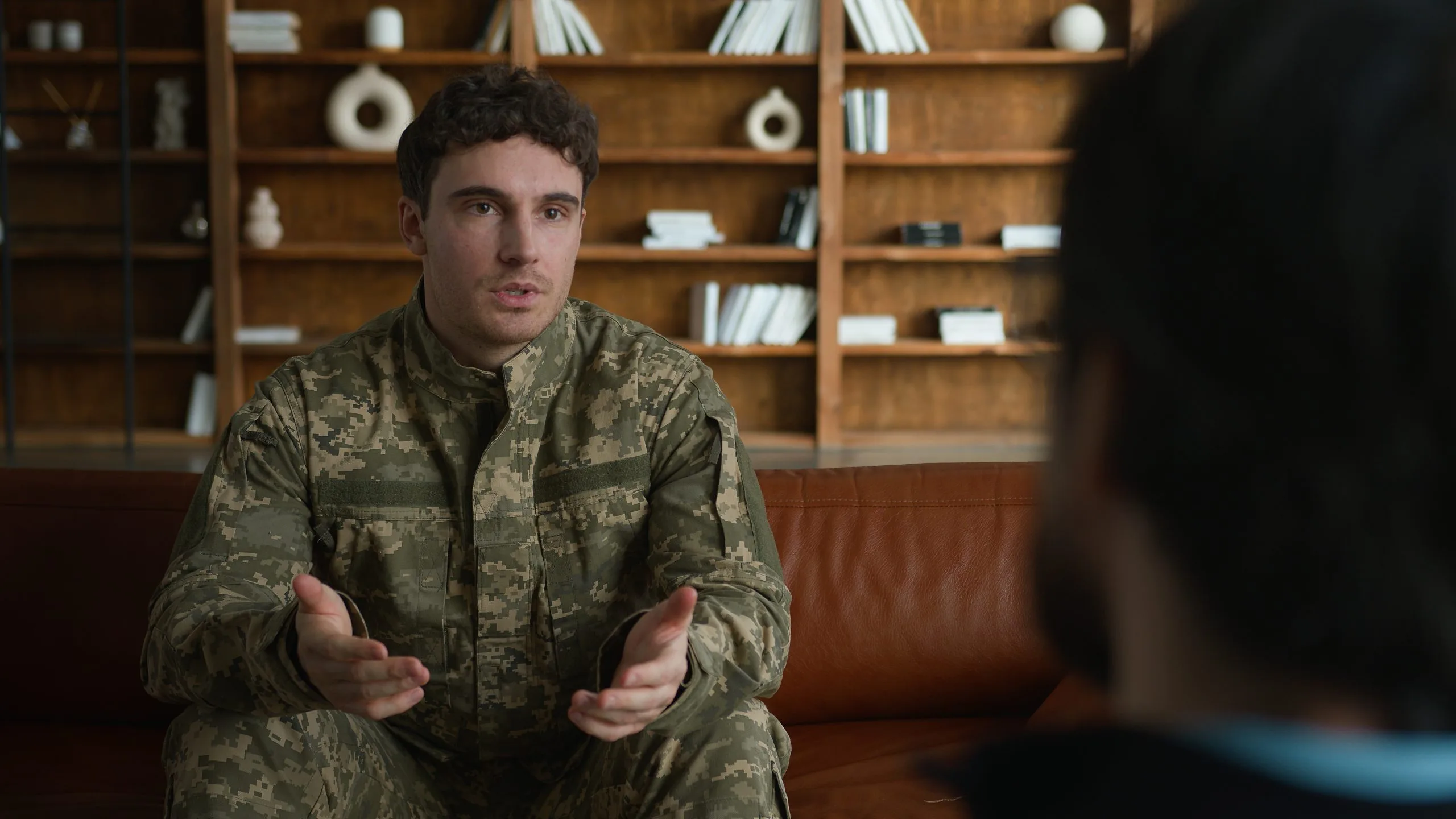 Service member in camouflage uniform gesturing while talking to counselor in office setting
