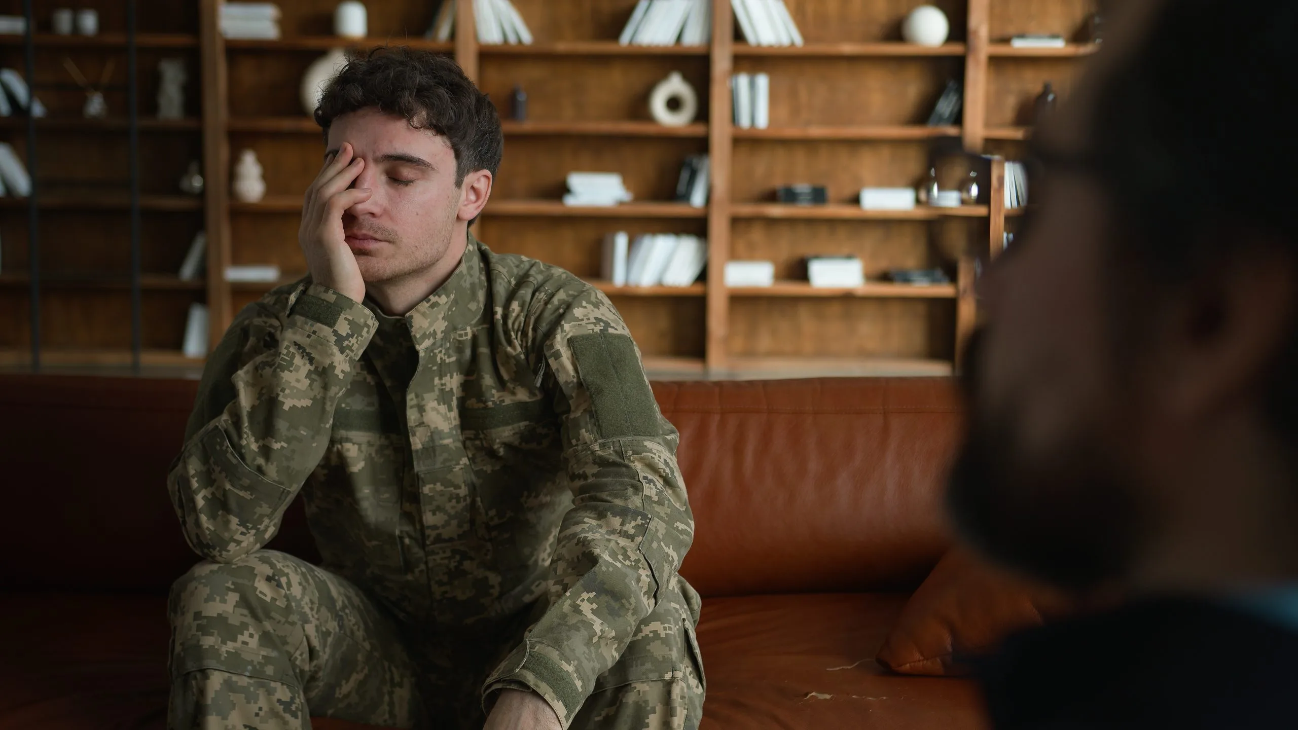 Service member in camouflage uniform with hand to face, appearing stressed during therapy session