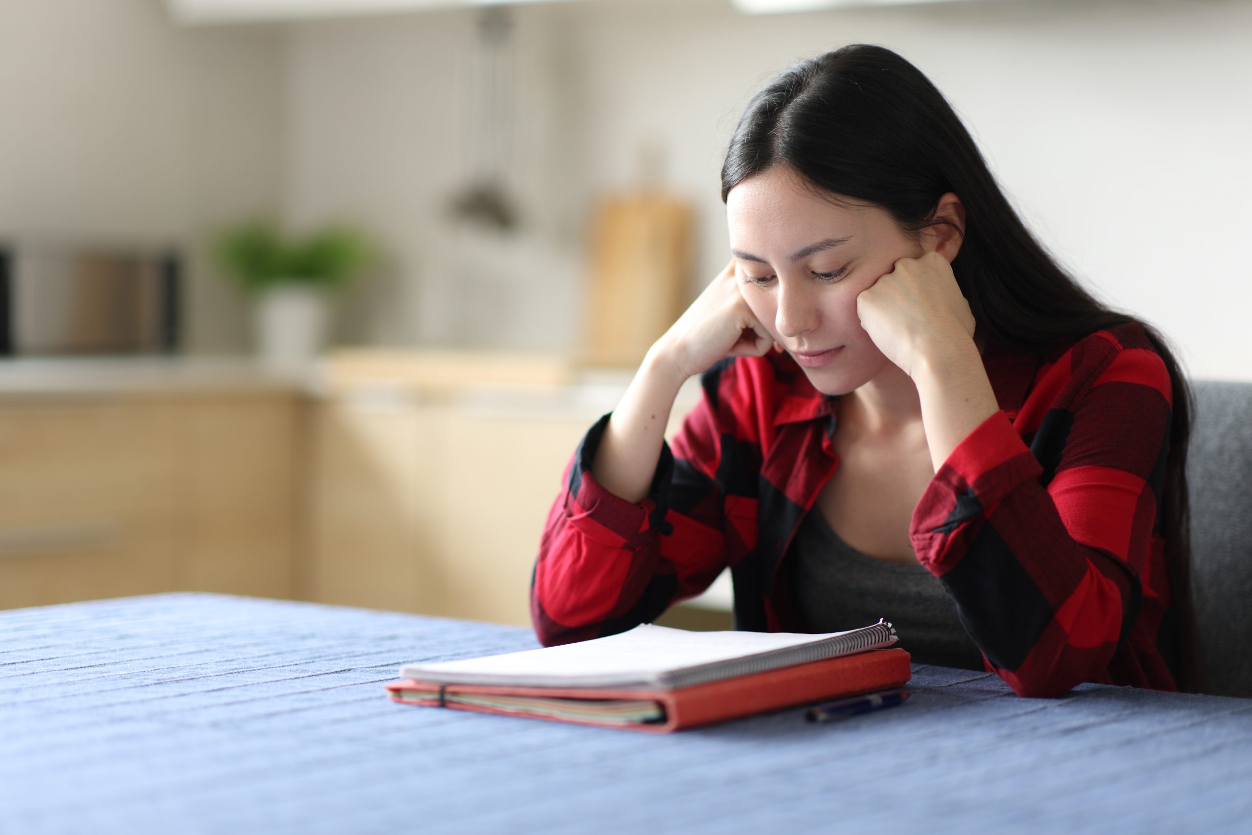 Woman in red plaid shirt reading notebook at table in kitchen