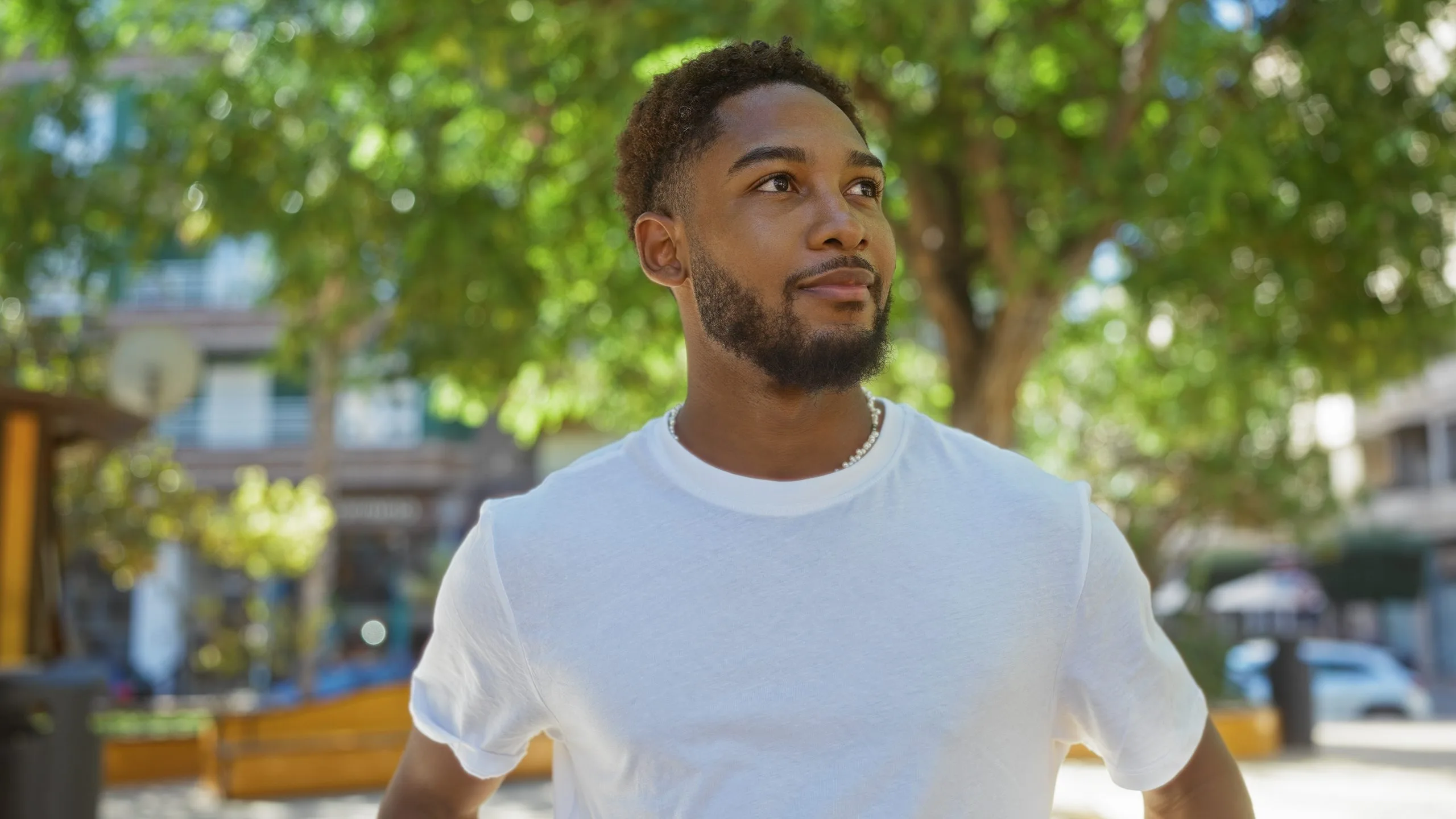 Young man in white t-shirt looking thoughtfully upward under tree canopy in urban plaza