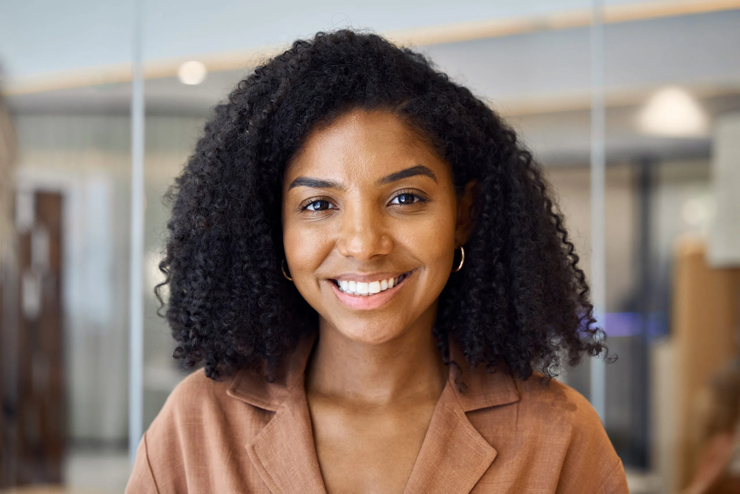 Woman smiling at camera in modern office hallway wearing rust-colored blazer