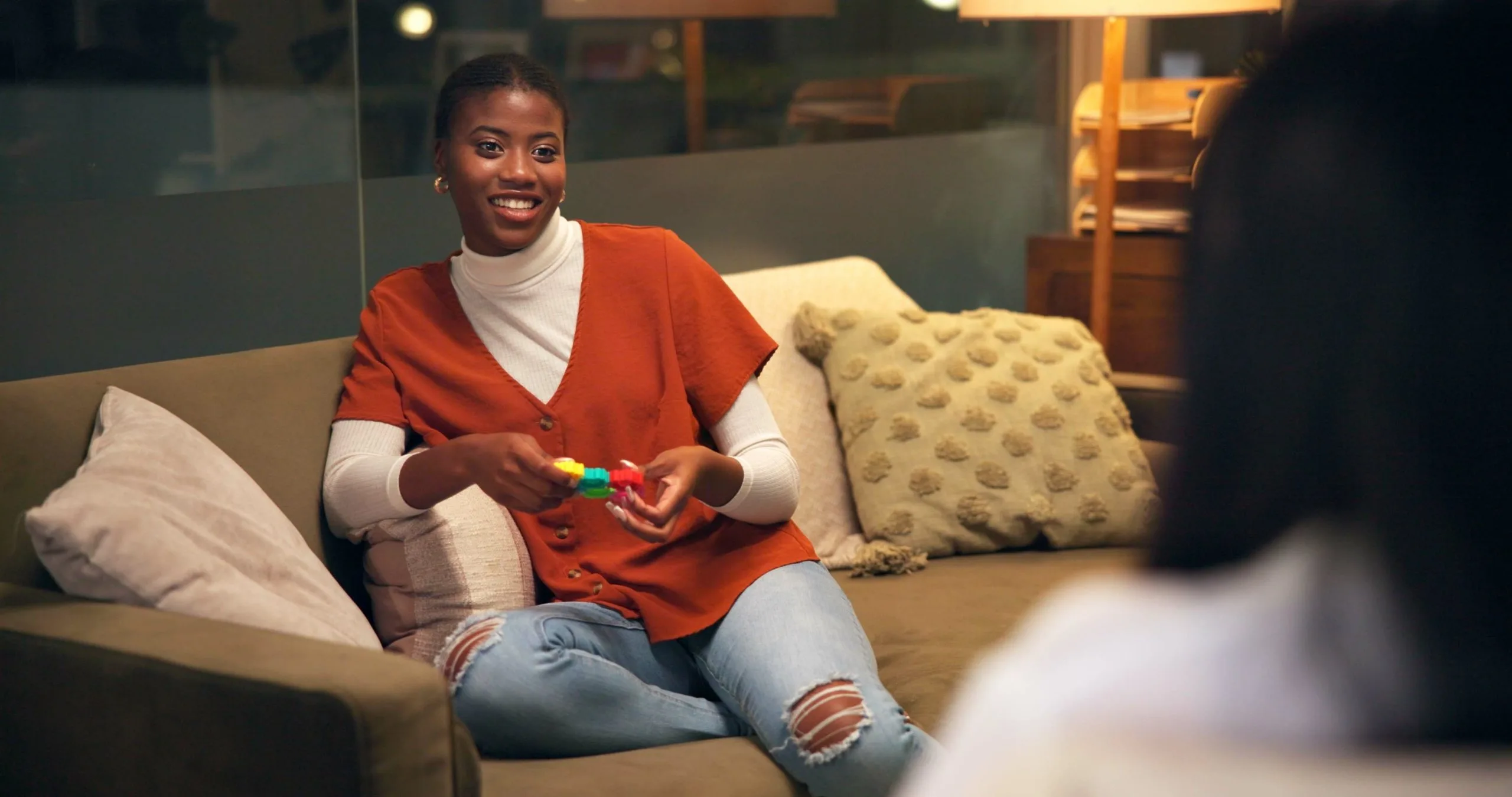 Woman in orange shirt smiling on a couch, holding a rainbow-colored stress relief toy during conversation