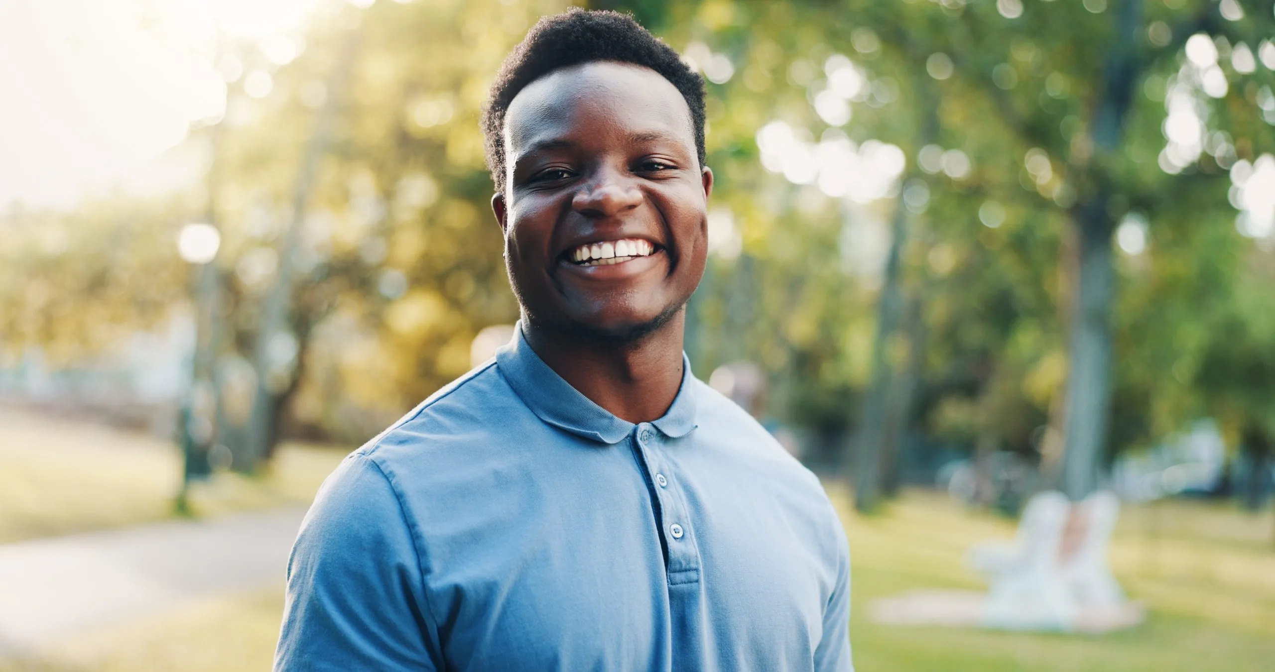 Smiling man in blue polo shirt standing outdoors with tree-lined path in background