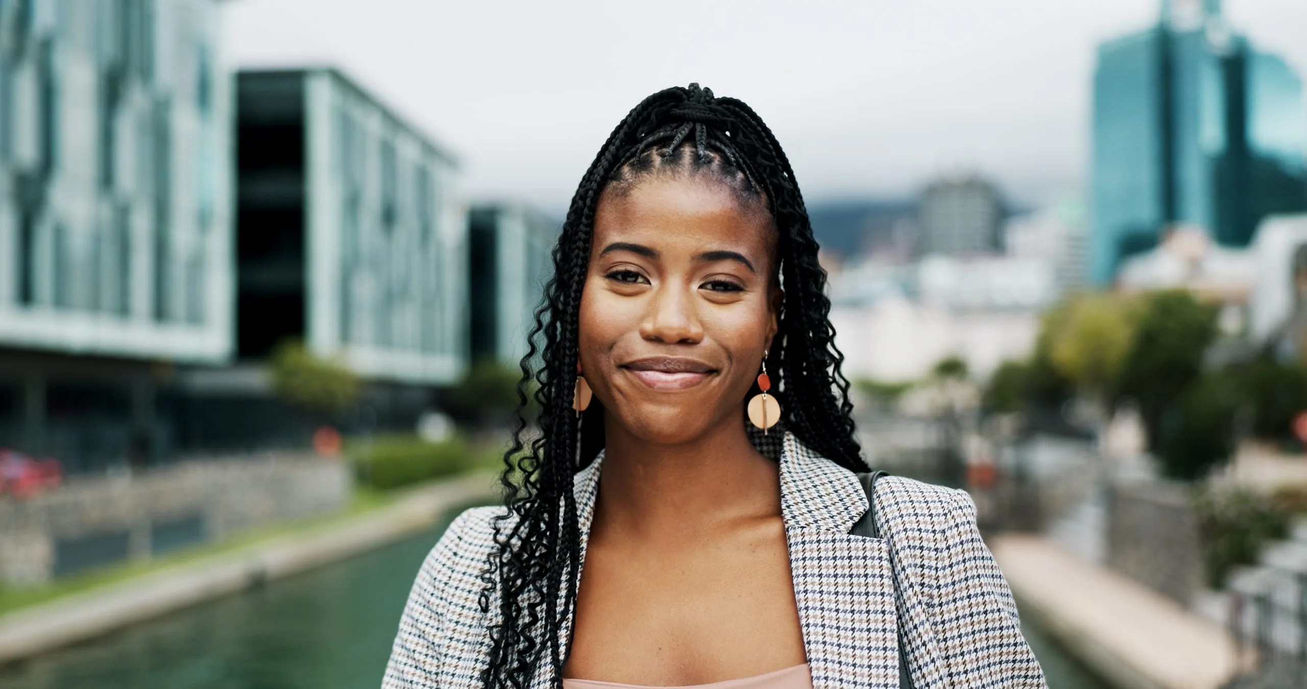 Woman with braided hair and pearl earrings smiling in front of urban skyline